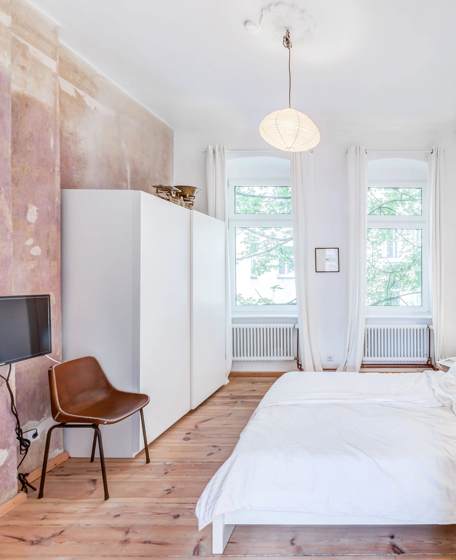 A minimalist bedroom featuring a white bed, a tall white wardrobe, and a brown leather chair against a distressed pink plaster wall.