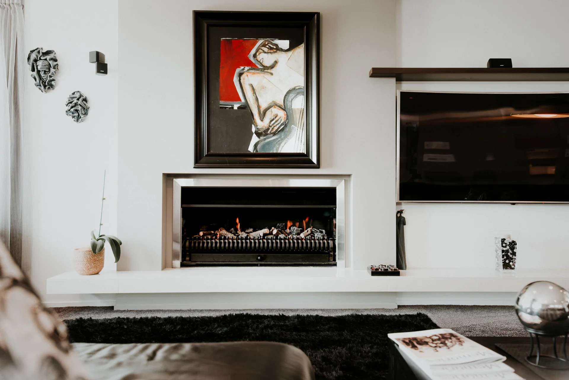 A modern off-white fireplace with active flames features a large framed abstract painting above and a flat-screen television to the right.
