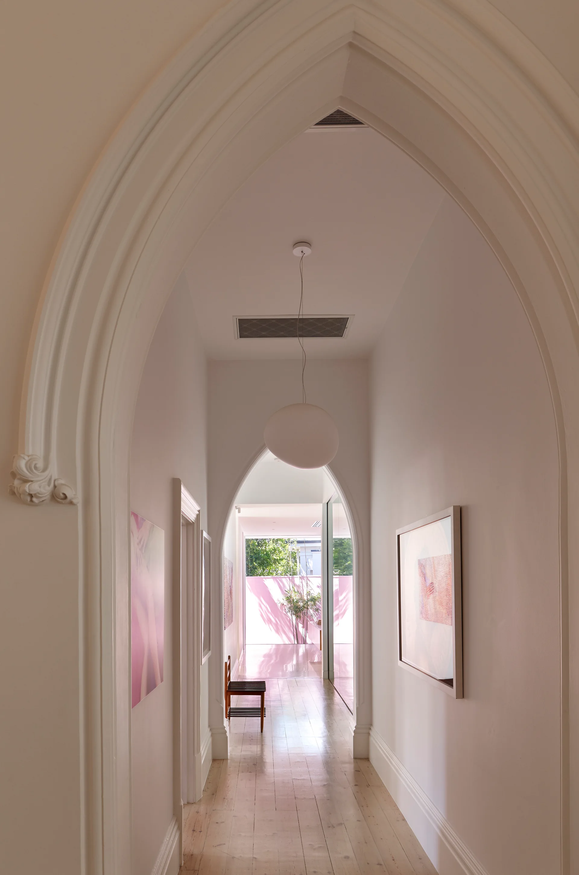 A white corridor features pointed gothic archways, light wood floorboards, framed art, and a round pendant light.