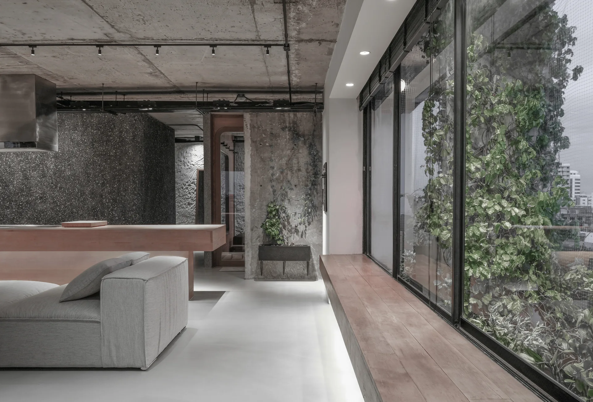 A modern minimalist room featuring a raw concrete ceiling, seamless light flooring, a grey sofa, and a long wooden bench under a window.