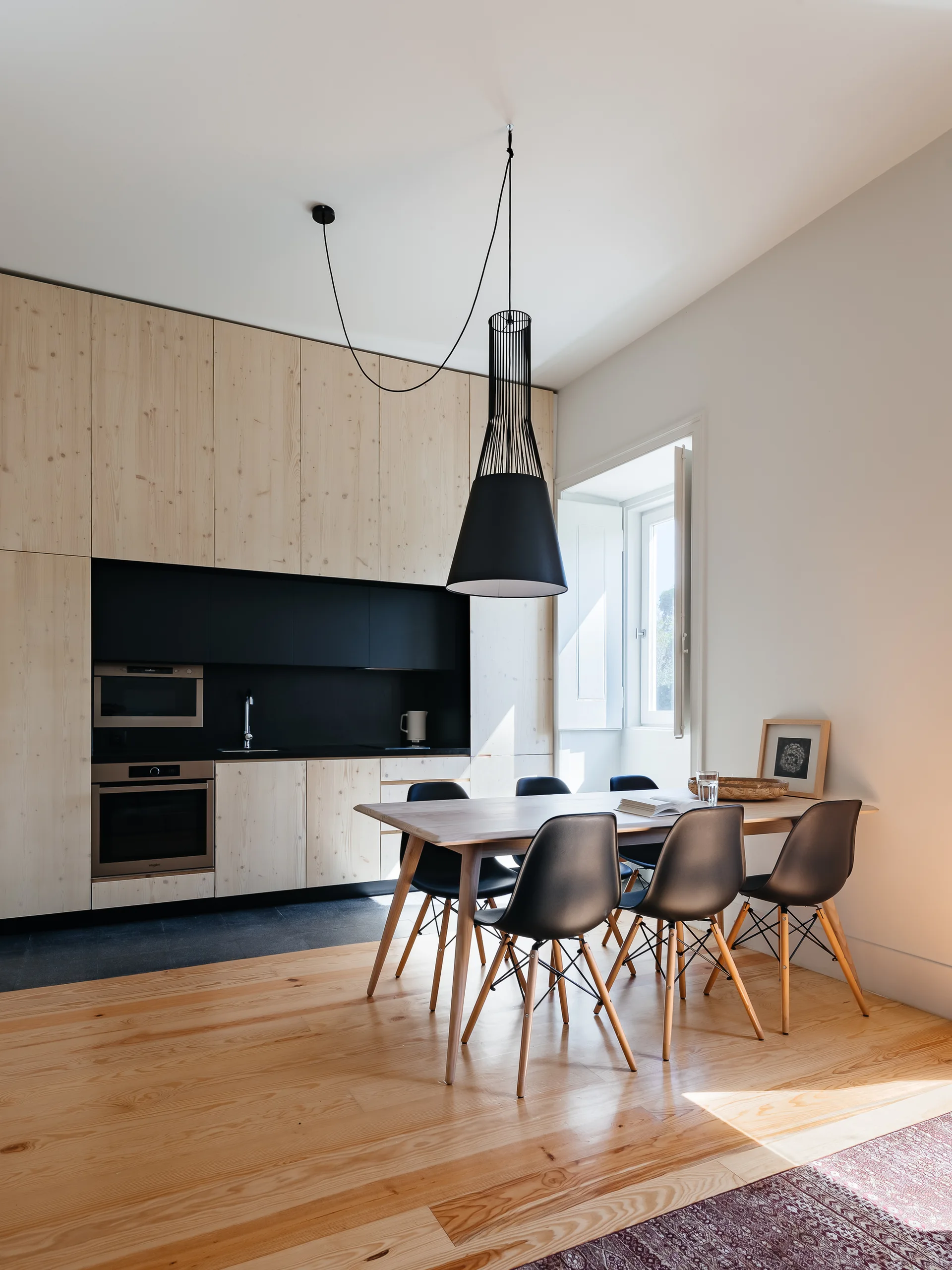 A minimalist kitchen and dining area featuring floor-to-ceiling light wood cabinetry, a black recessed cooking niche, and black dining chairs.