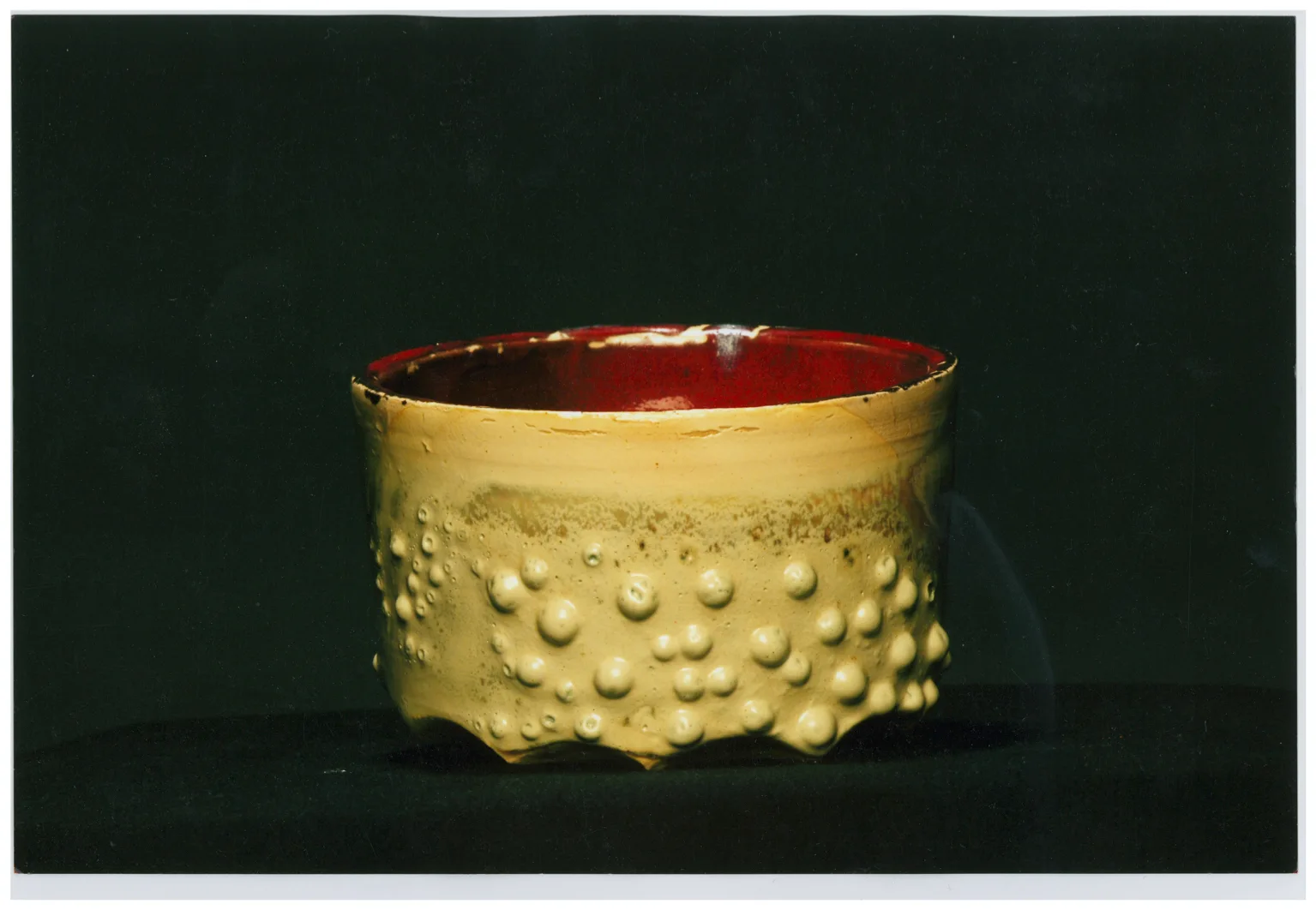A handcrafted ceramic bowl featuring a bubbly beige exterior and a glossy red interior glaze displayed against a solid dark backdrop.