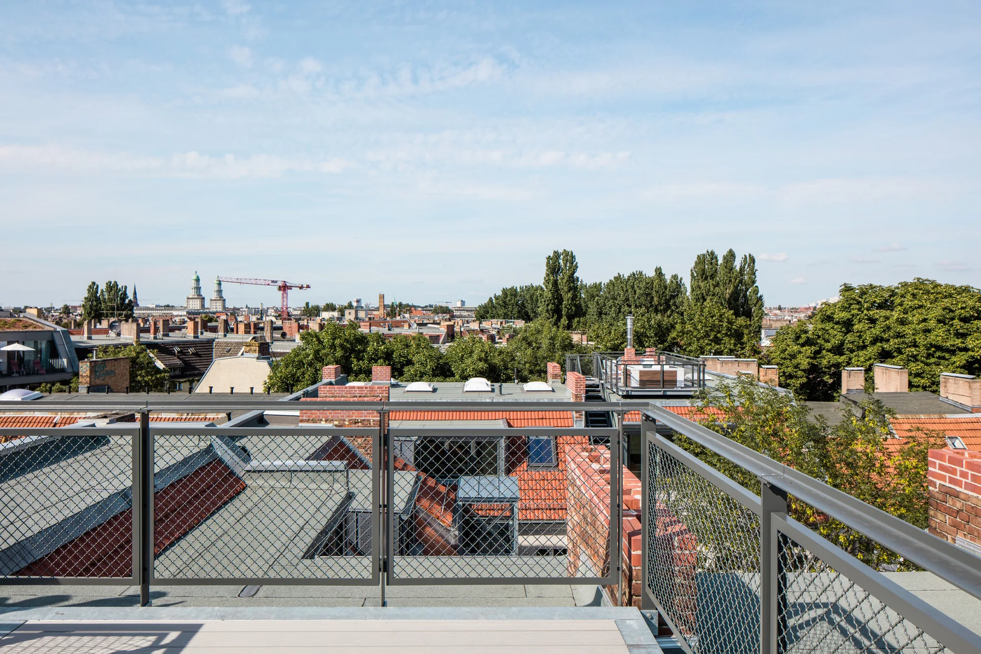 An elevated rooftop terrace features a grey metal railing with wire mesh panels looking out over a city skyline with terracotta roofs under a blue sky.