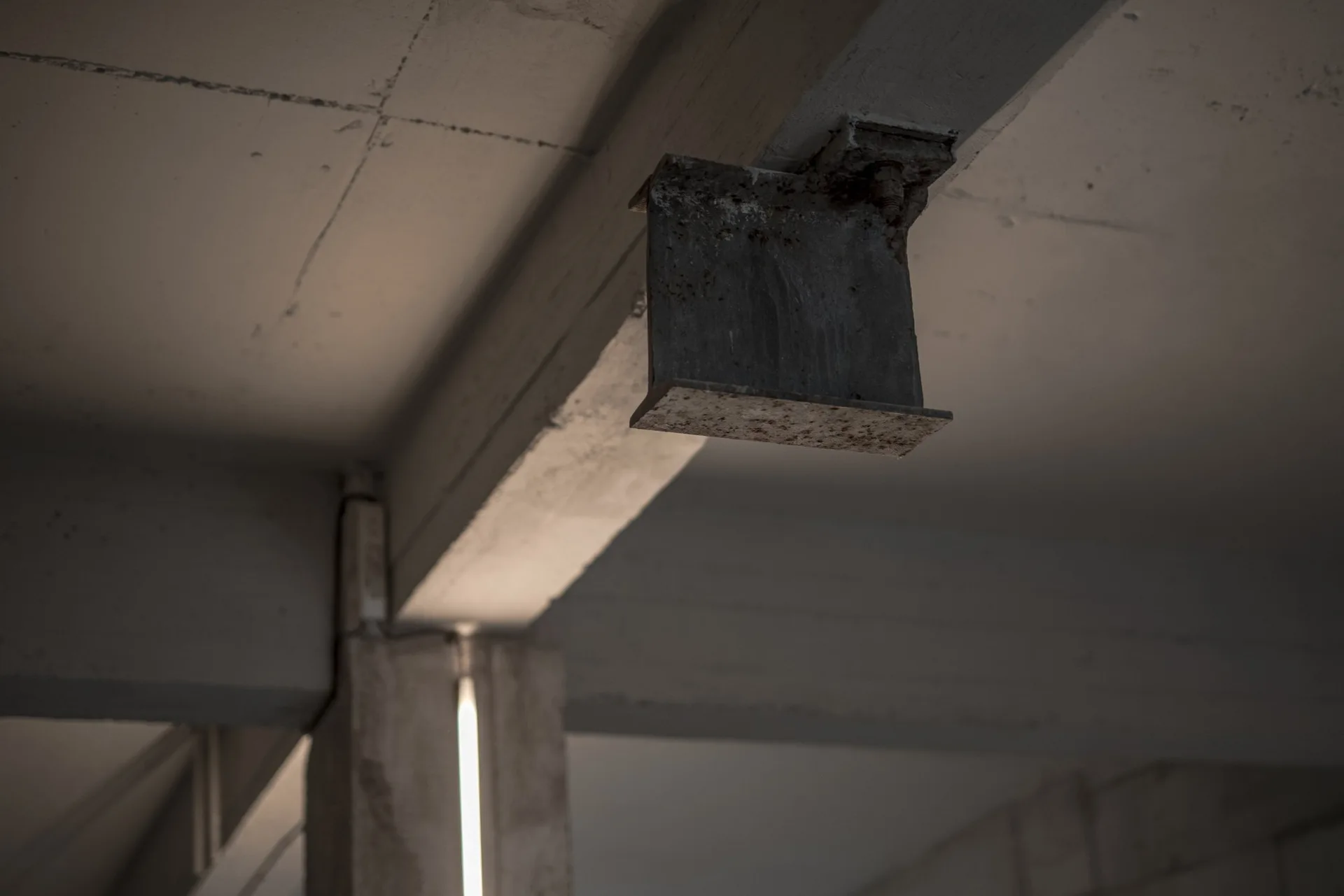A close-up of a structural grey concrete ceiling beam with an attached rusted metal bracket in an industrial interior.
