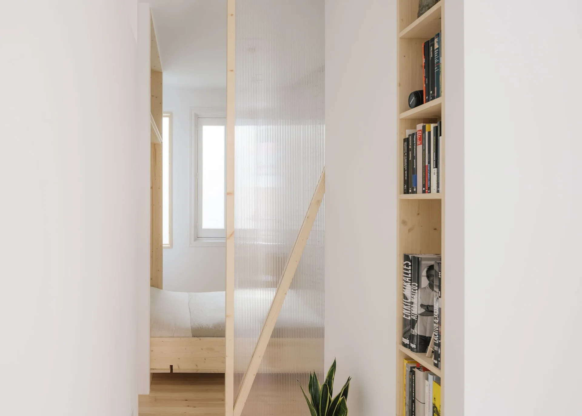 A fluted glass partition divides a narrow white hallway with built-in wooden shelves from a minimalist bedroom area.