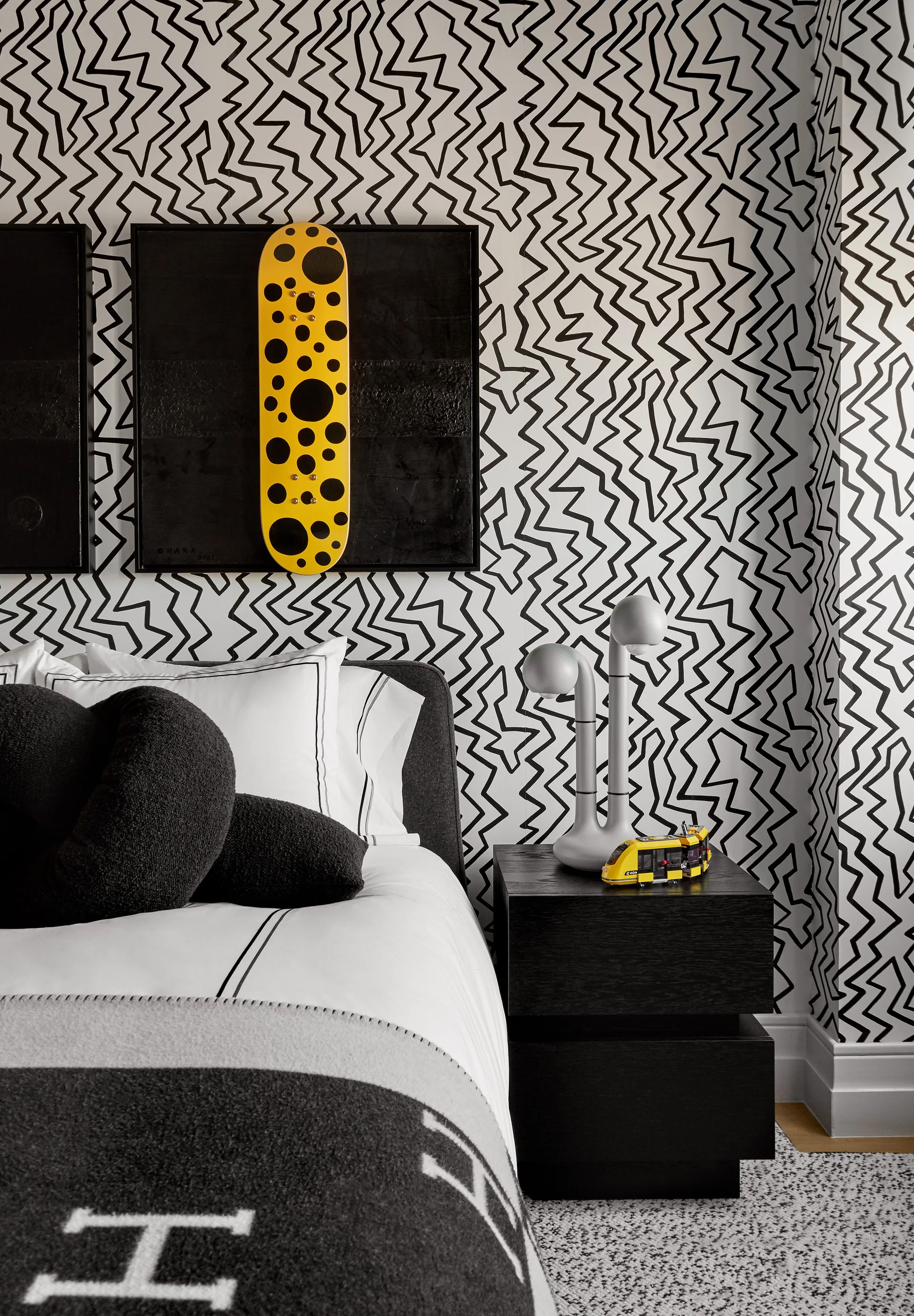 A bedroom features bold black and white zig-zag wallpaper, a dark upholstered bed, and a black wood nightstand with a grey sculptural lamp.