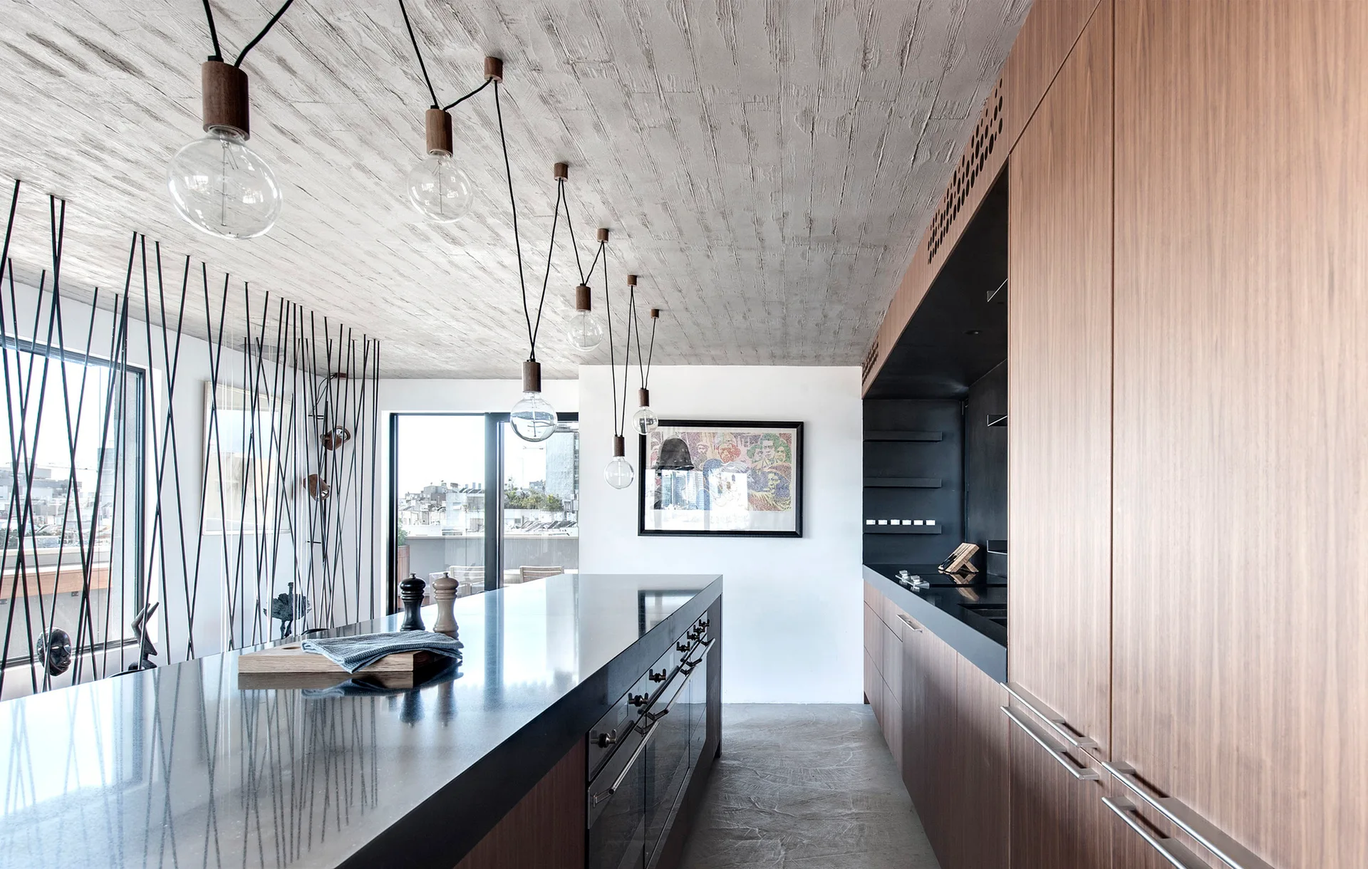 A modern kitchen with a long black island, warm wood cabinetry, a textured concrete ceiling, and hanging exposed bulb pendant lights.