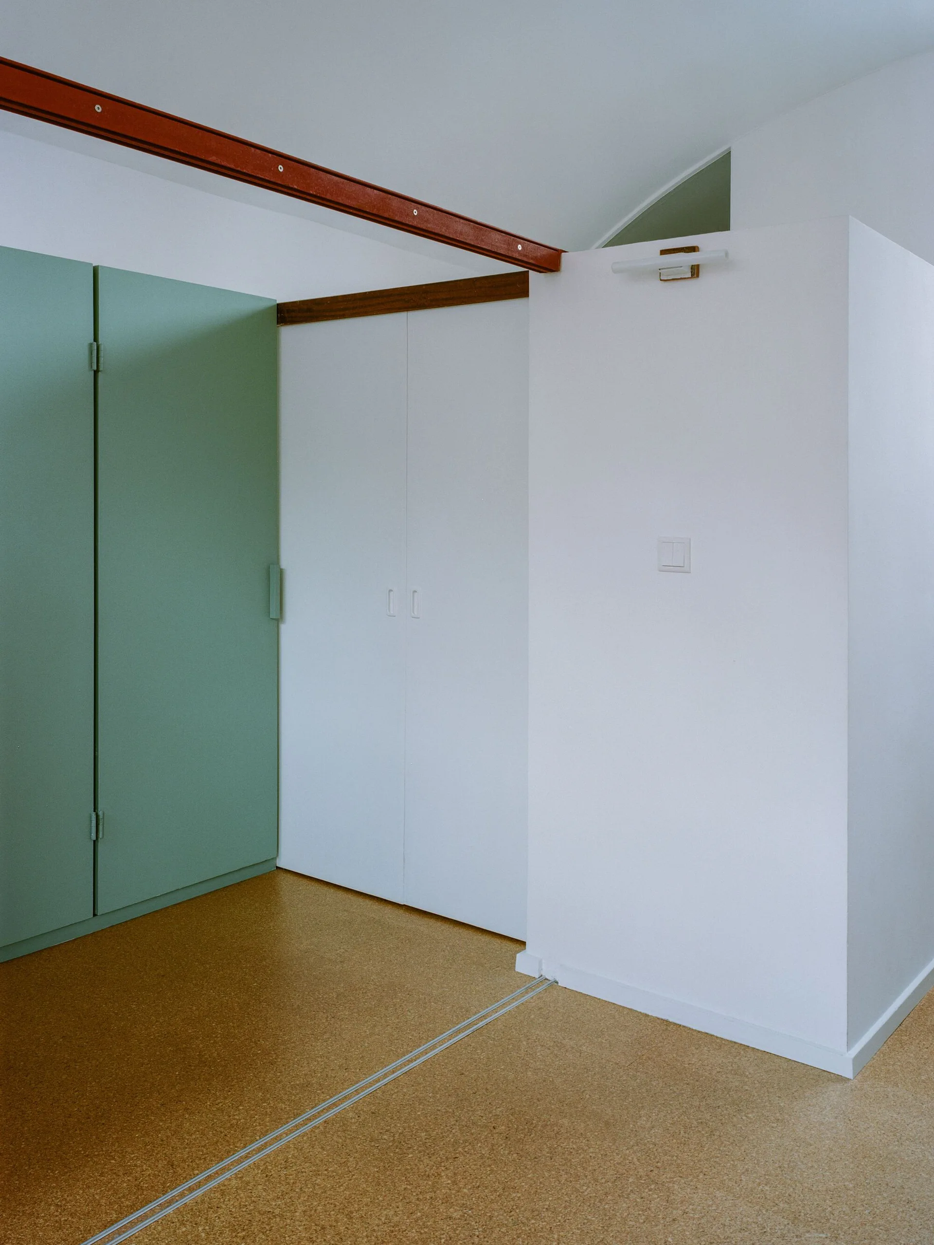 A minimalist interior features sage green and white built-in wardrobes, a warm cork floor, and an exposed orange metal ceiling beam.
