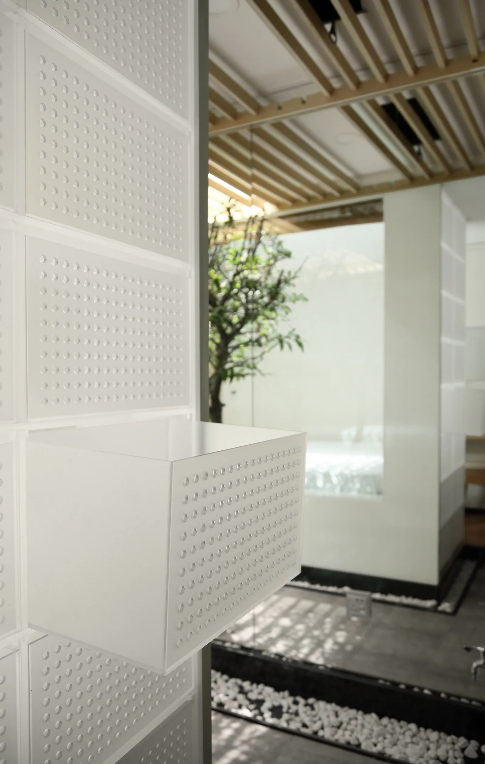 A close-up of white perforated wall panels with an attached storage box near a slatted wood ceiling and indoor pebble garden.