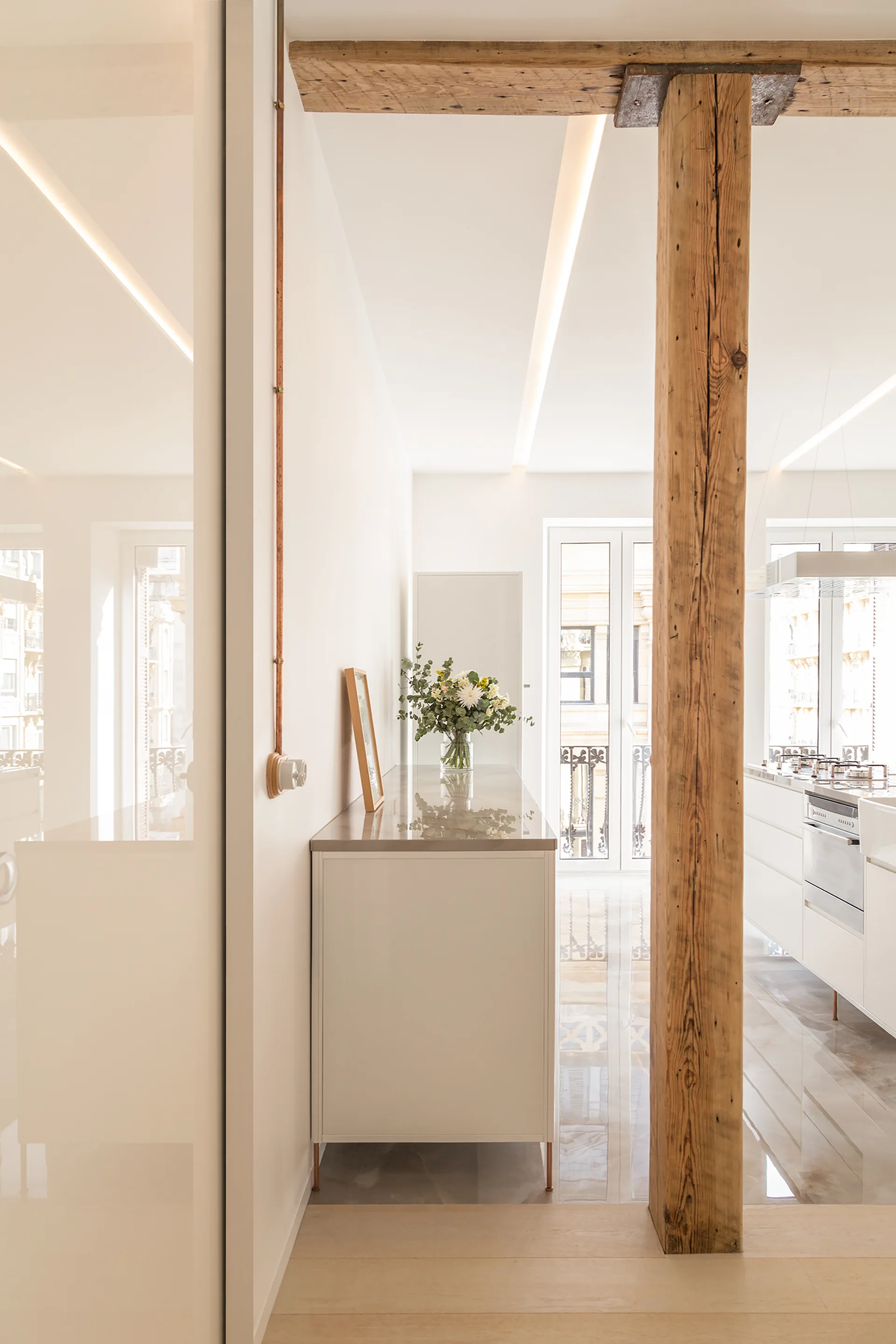 A minimalist white kitchen with shiny light-colored flooring, recessed linear ceiling lights, and a weathered vertical timber support beam.