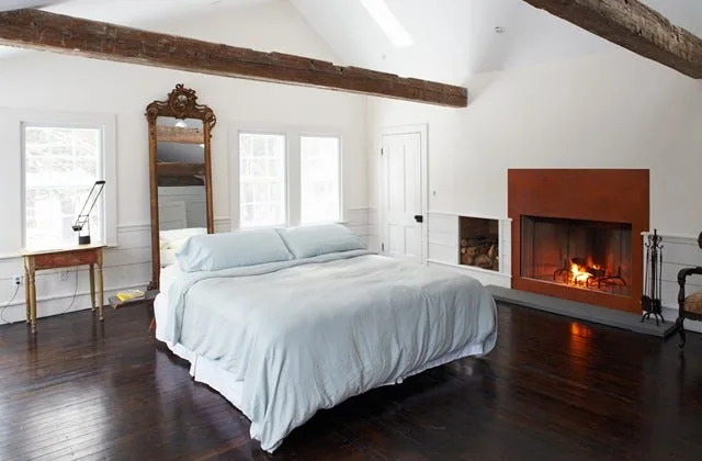 A rustic bedroom featuring dark wood floors, white wainscoted walls, an exposed timber ceiling beam, a fireplace, and a bed with light blue bedding.