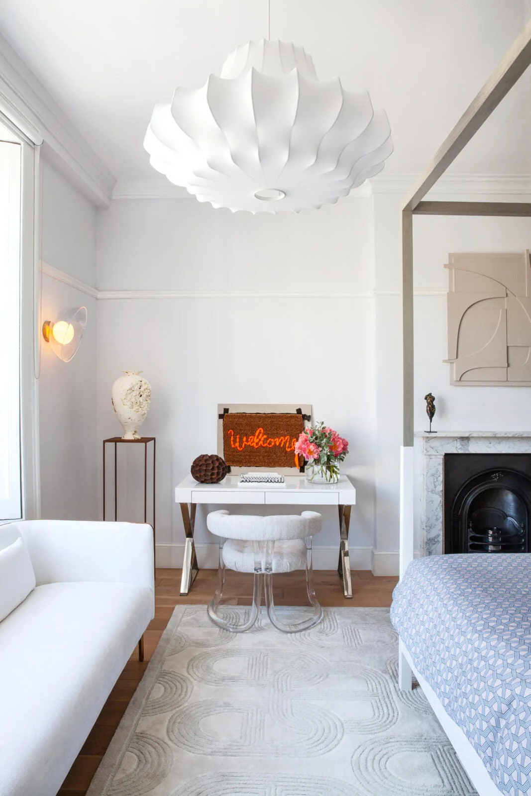 A bright bedroom featuring a white writing desk, a curved chair with clear acrylic legs, a marble fireplace, and a large pleated pendant light.