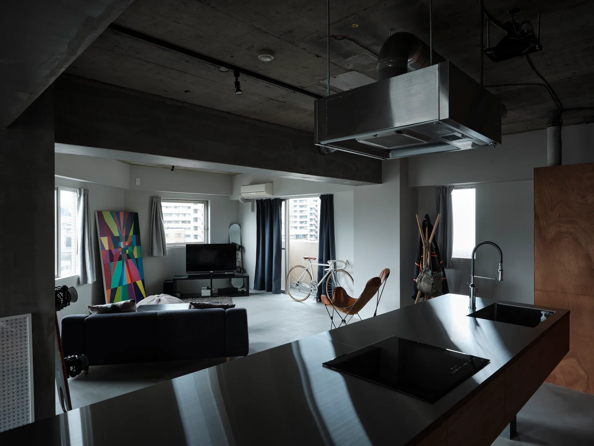 An industrial loft apartment with a brushed metal kitchen island in the foreground and a living area with an exposed concrete ceiling.