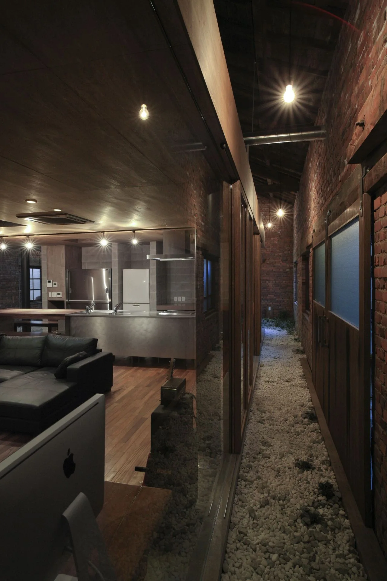 An industrial loft interior features a black leather sofa and kitchen adjacent to a narrow, indoor gravel walkway lined with exposed brick walls.