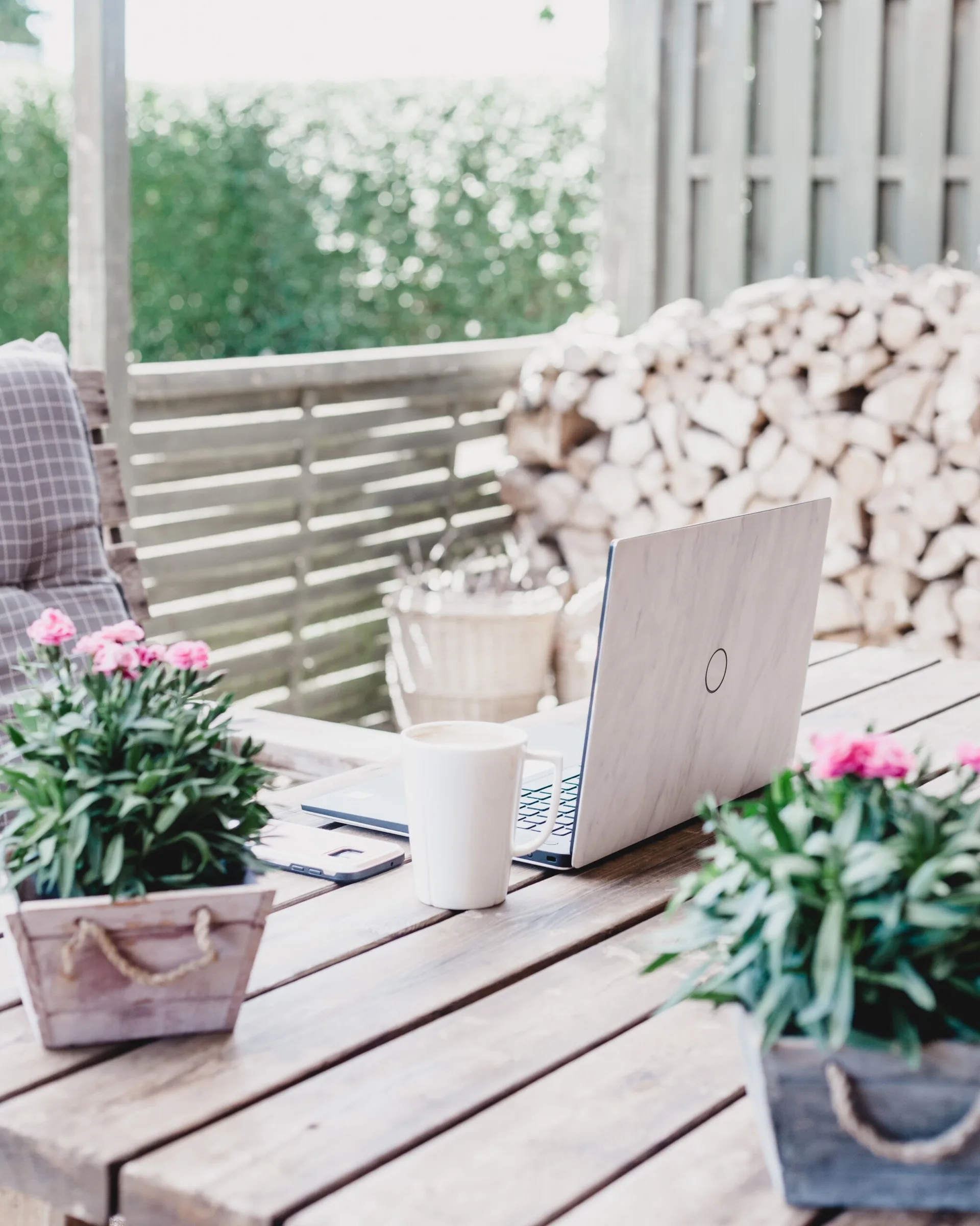 A laptop, white mug, and rustic crate planters with pink flowers sit on a wooden patio table, with stacked firewood in the background.