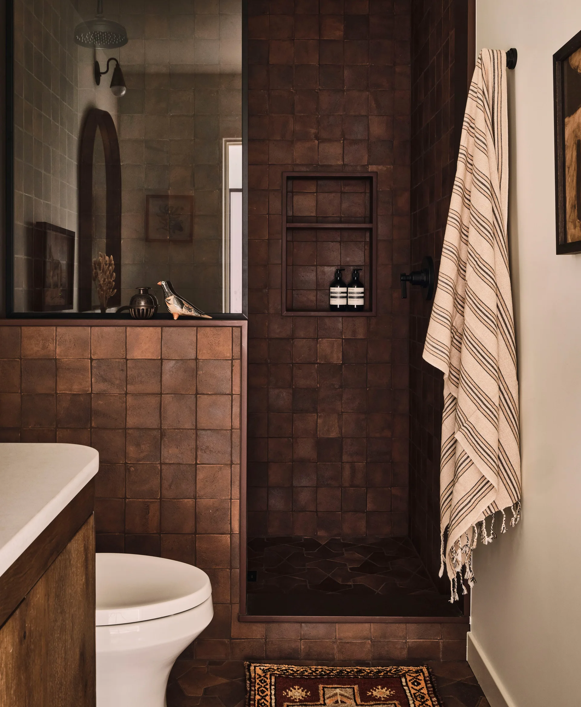 A modern bathroom features a walk-in shower lined with dark brown square tiles, a white toilet, and a hanging striped towel.
