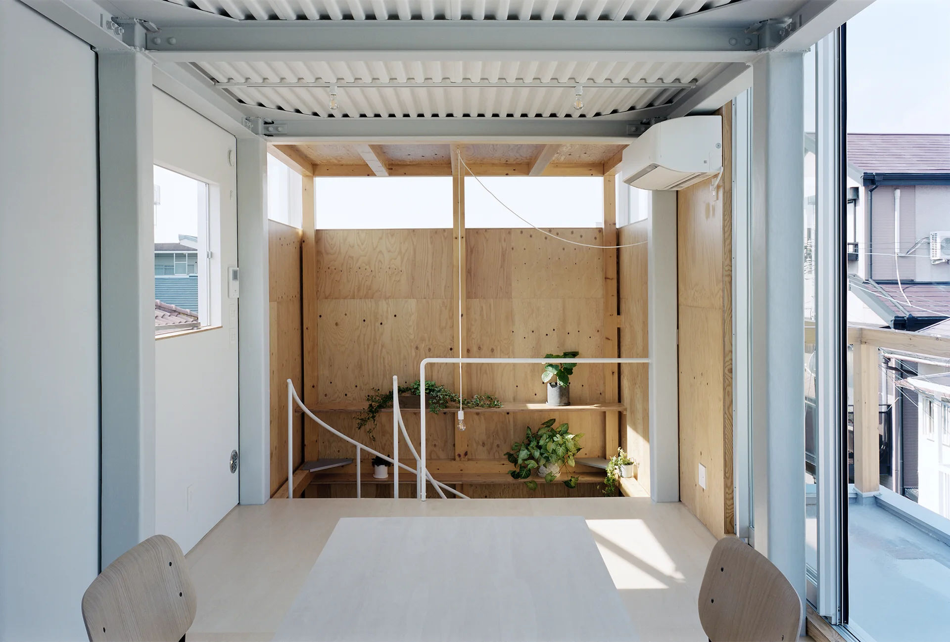 A minimalist dining area features a light wood table, a corrugated steel ceiling, white steel beams, and plywood walls with built-in shelves.