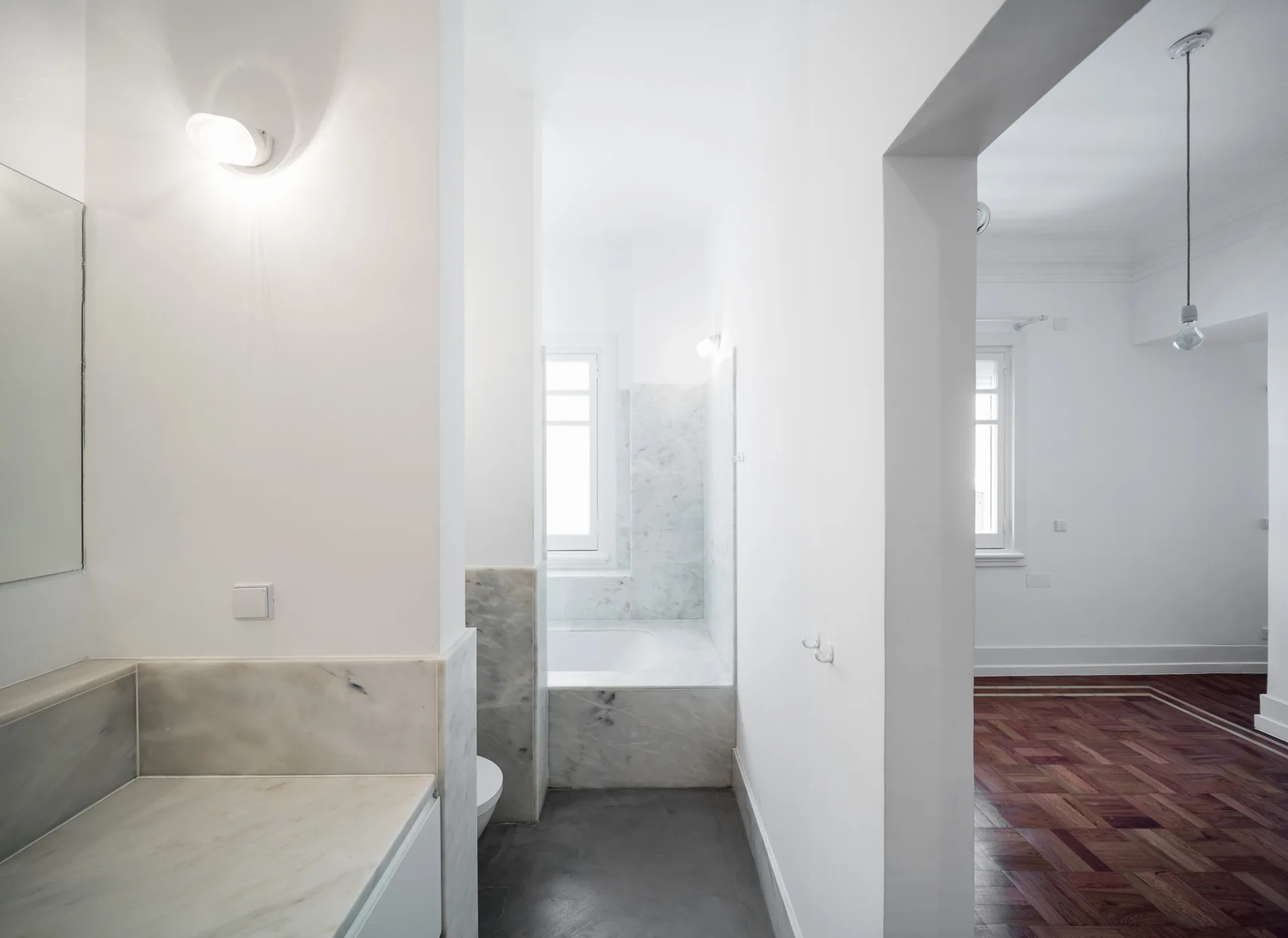 A minimalist white bathroom with a marble-clad tub next to a hallway that opens to a room with dark wood parquet flooring.