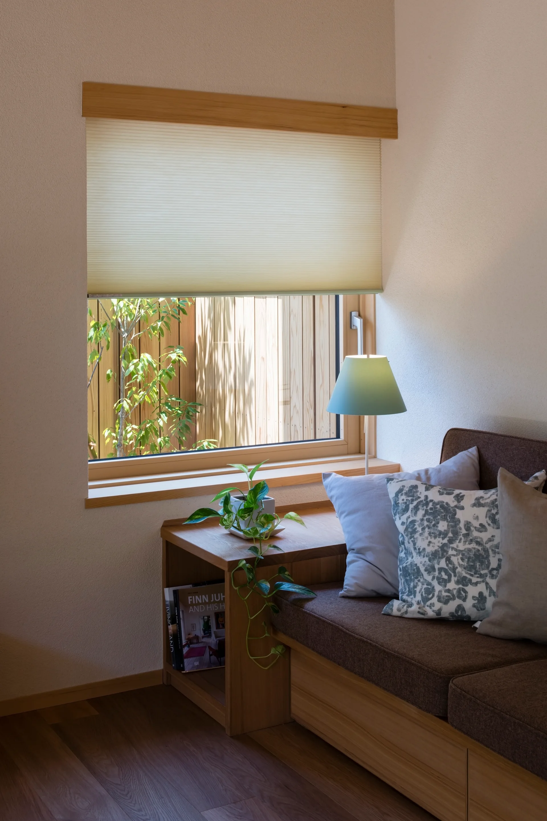 A built-in wooden seating bench with brown cushions and throw pillows sits next to a window with a cellular shade and a small table lamp.