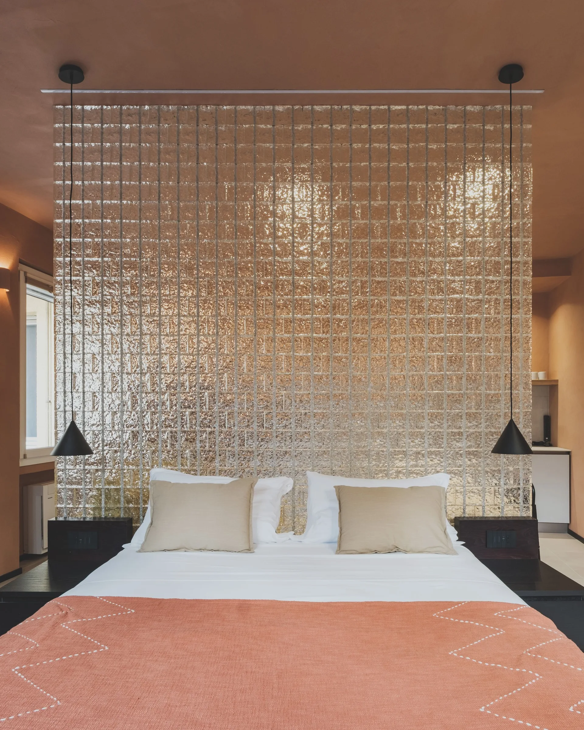 A bedroom with an orange wall and ceiling features a bed set against a textured glass block wall with black pendant lights.