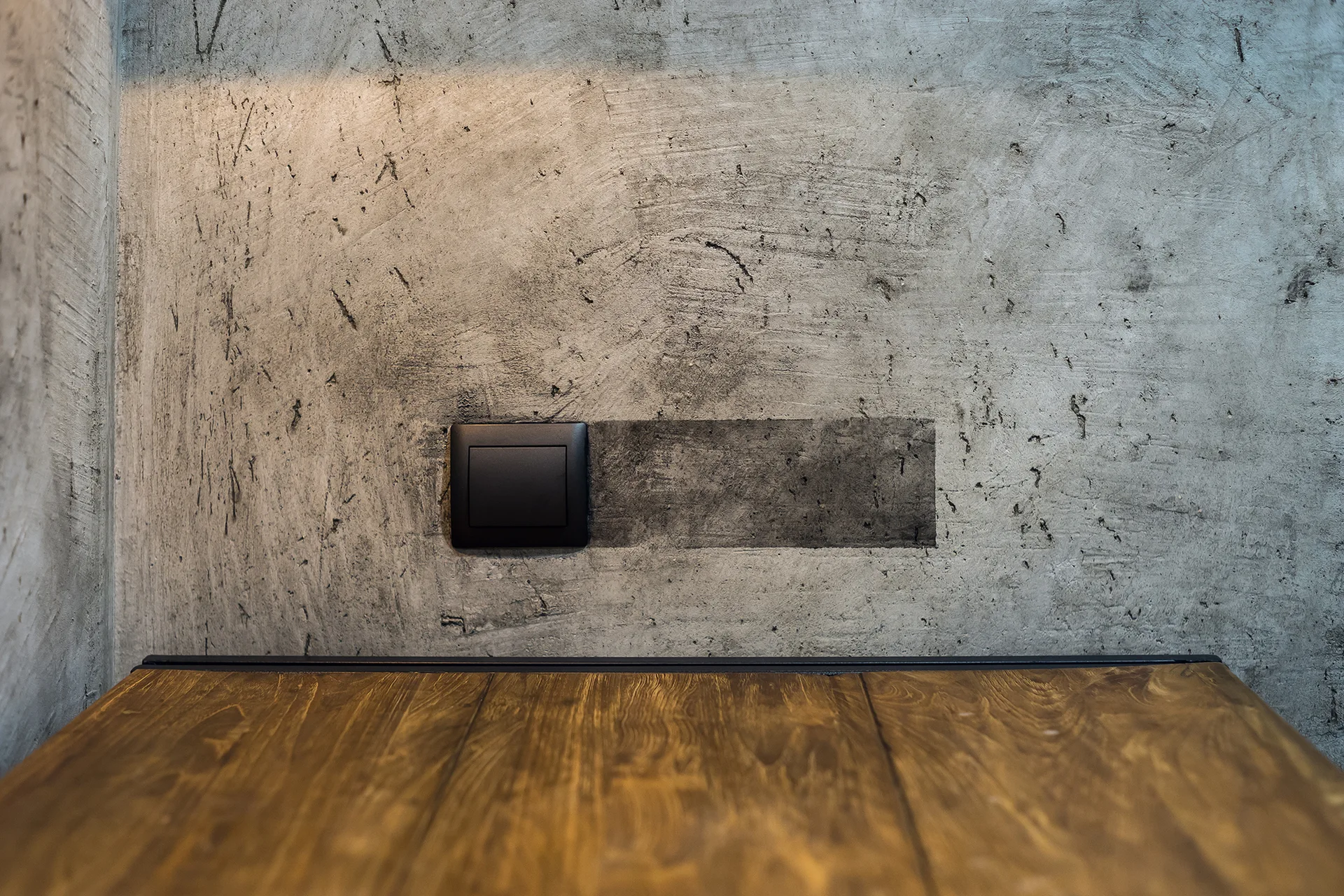 A close-up of a matte black square electrical switch on a textured grey concrete wall above a warm brown wood surface.