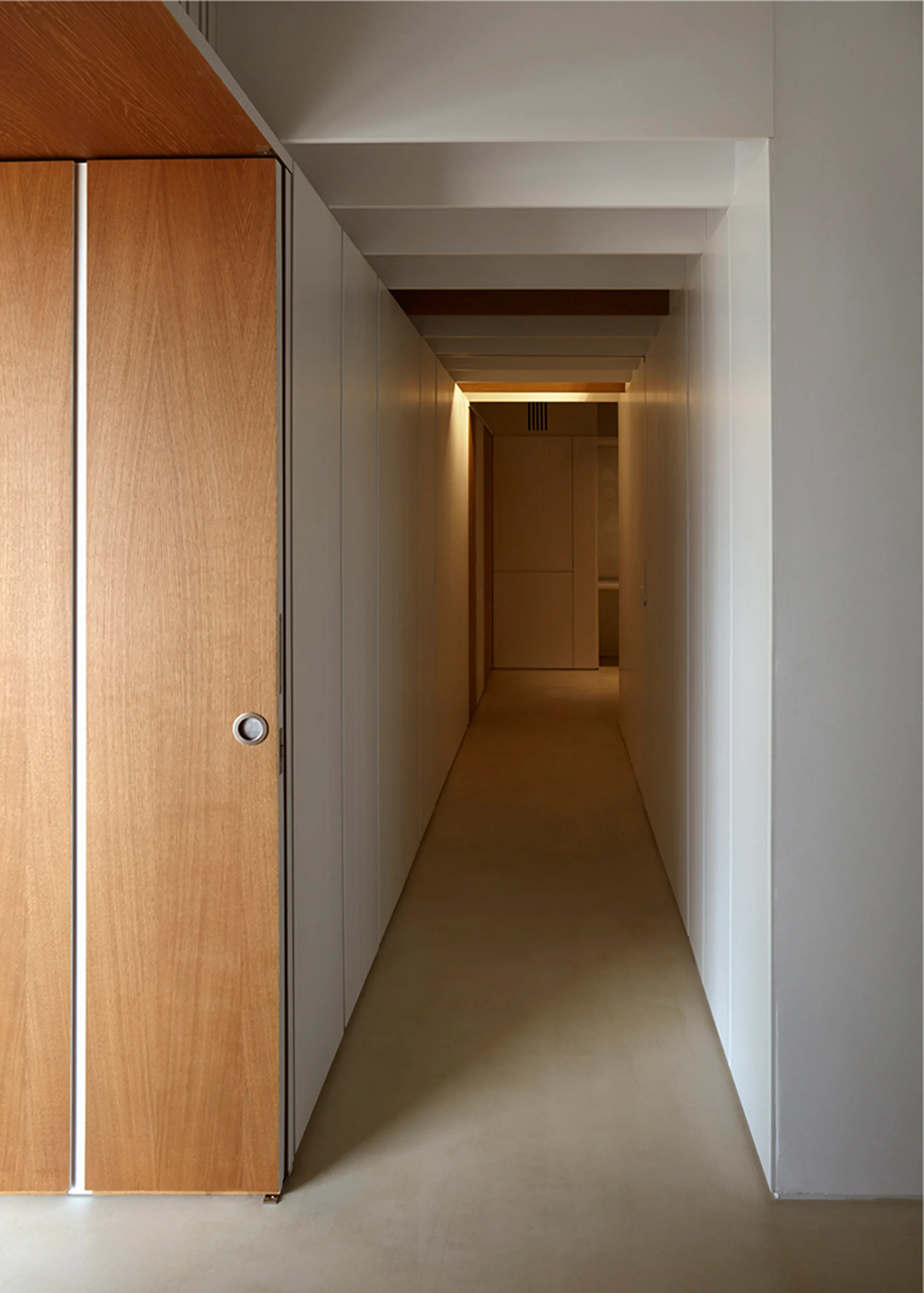 A minimalist hallway features vertical white wall paneling, a light wood door, and structural ceiling beams leading to an illuminated corner.