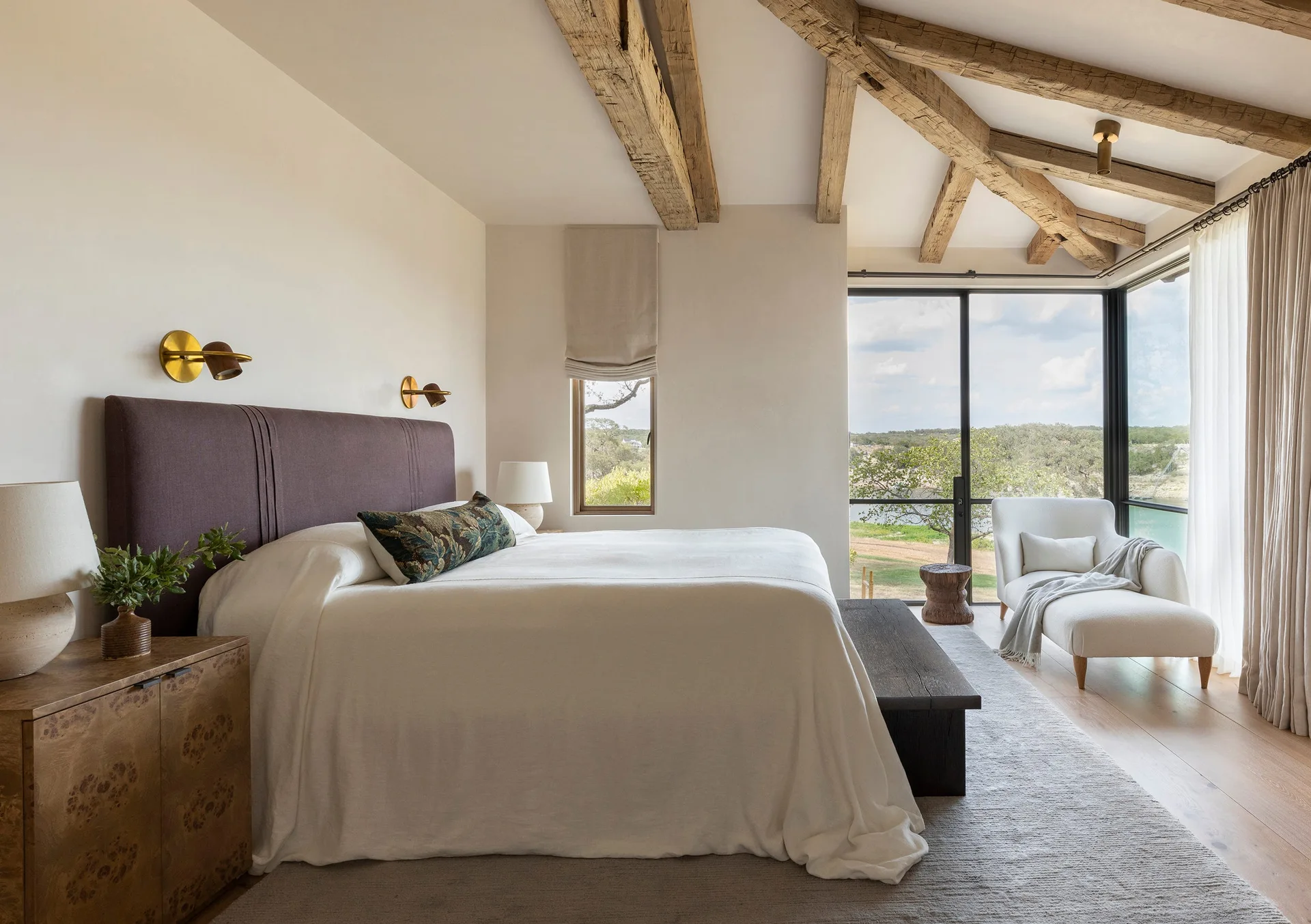 A neutral bedroom features rustic wood ceiling beams, an upholstered dark headboard, a wooden footbench, and a scenic window view.