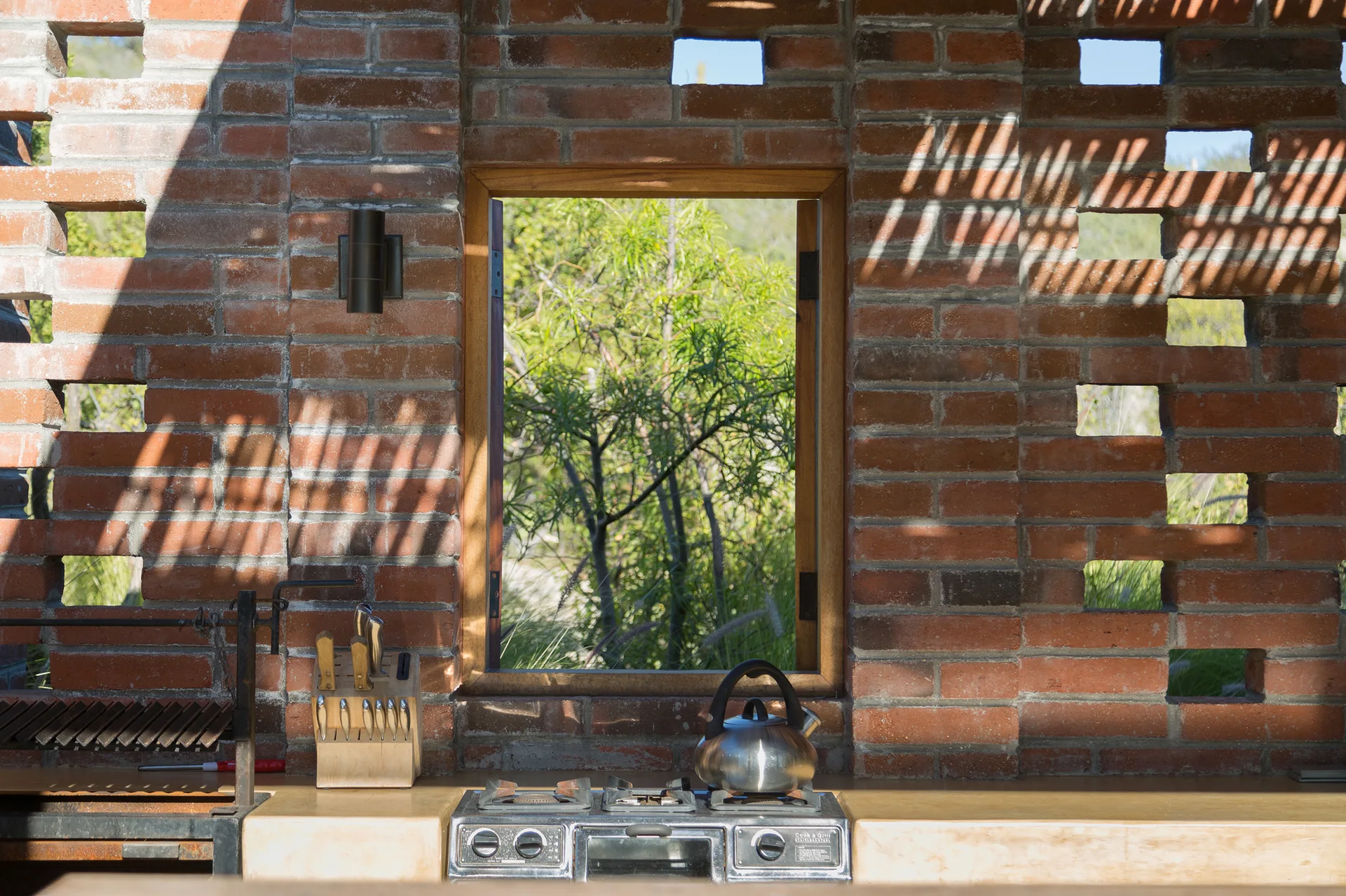 An outdoor kitchen features a built-in stove on a light concrete counter against an open-grid terracotta brick screen wall with a central wood-framed window.
