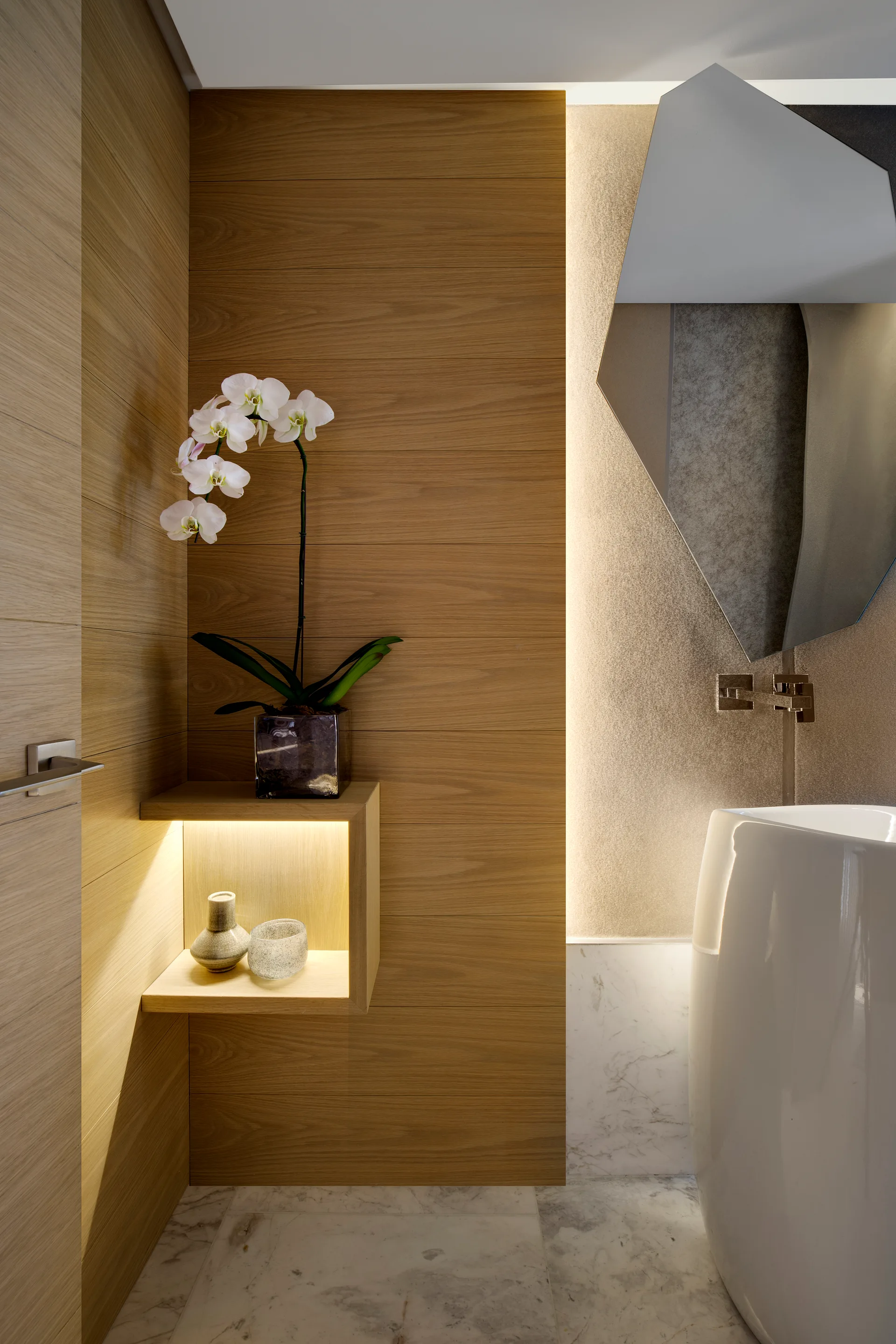 A contemporary powder room features horizontal wood paneling, a white pedestal sink, a geometric wall mirror, and an illuminated floating niche shelf.