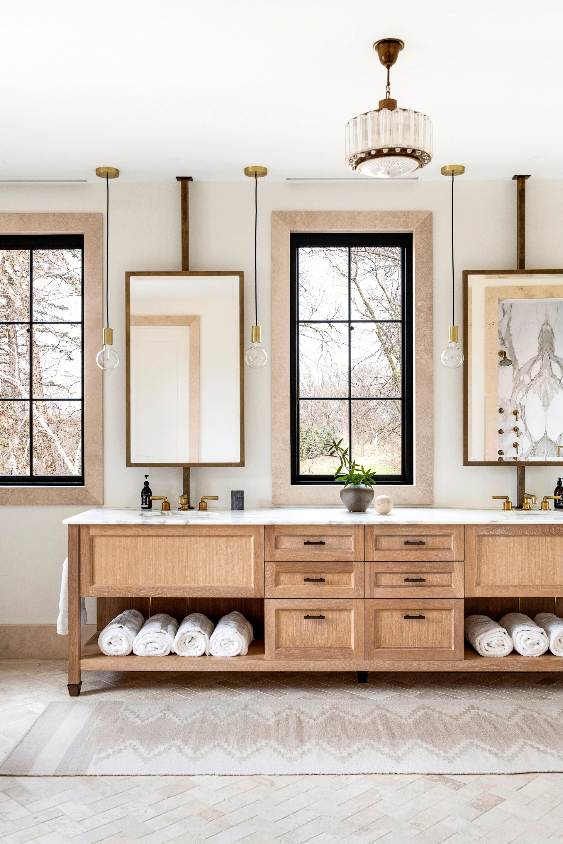 A light oak double vanity features marble countertops and hanging mirrors situated between stone-cased windows with black frames.