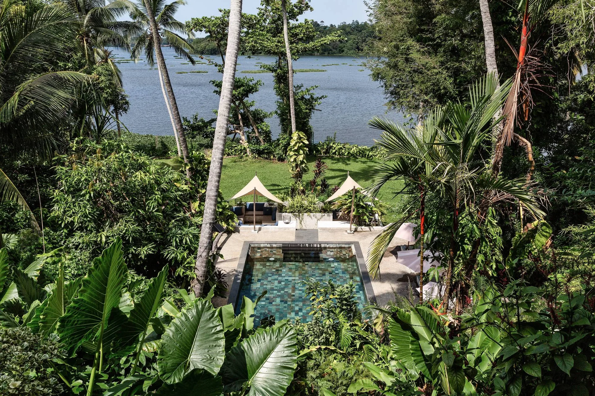 A rectangular tiled swimming pool sits in a lush tropical garden with beige lounge umbrellas and a scenic waterfront view in the background.