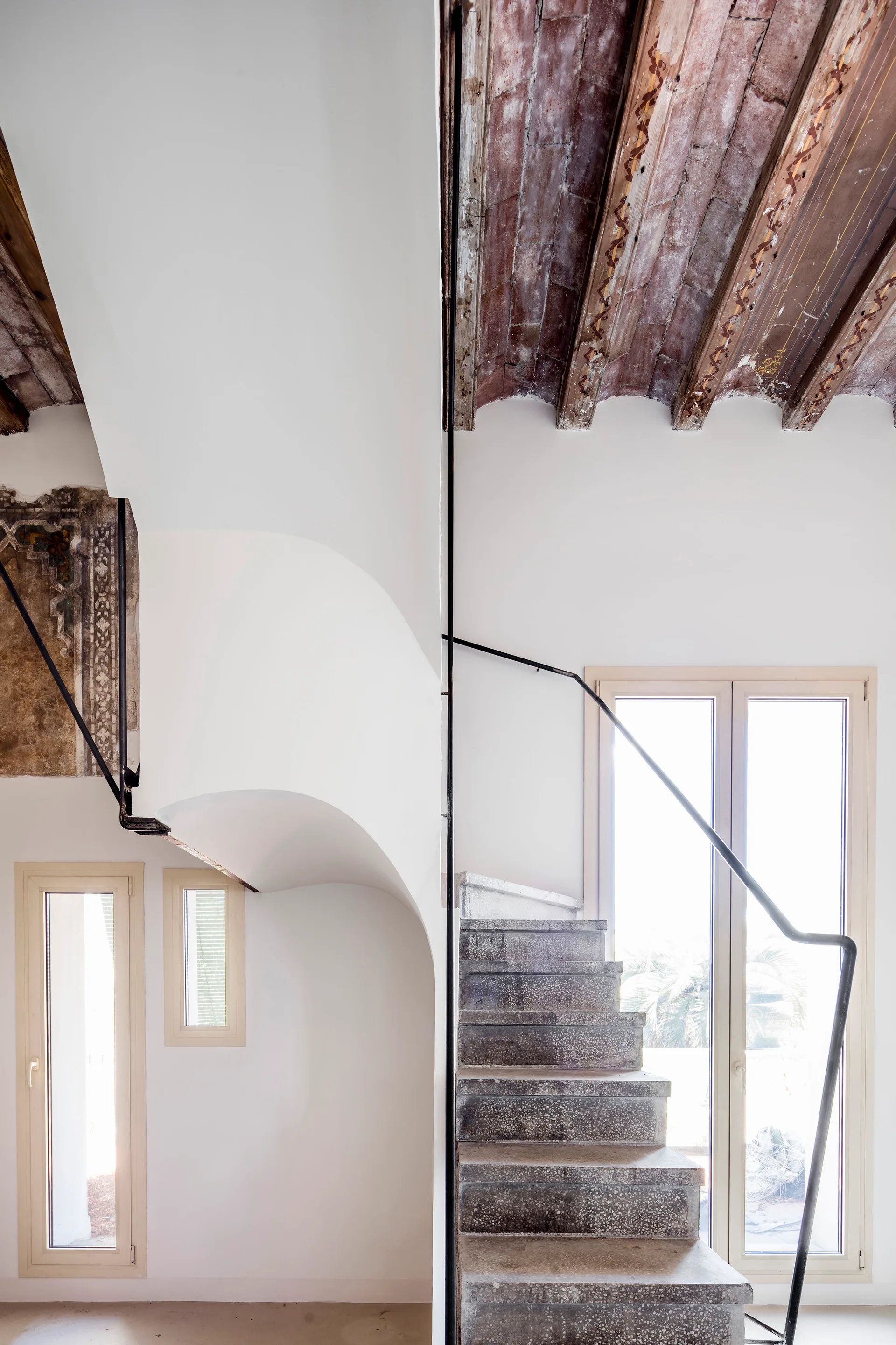 A stone staircase with a black metal railing stands next to a curved white wall beneath an exposed wooden beam ceiling.