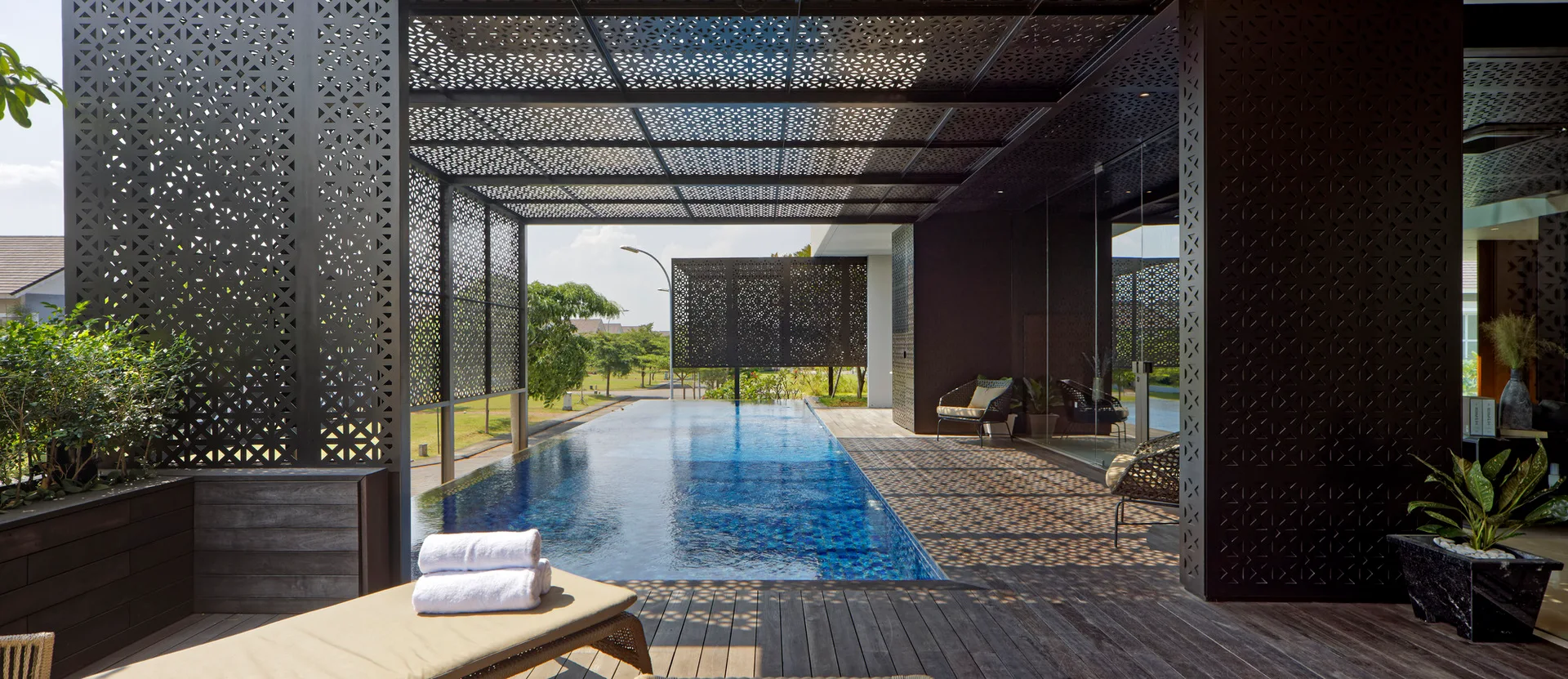 A rectangular swimming pool is flanked by a wooden deck under a black laser-cut metal canopy that casts geometric shadows.