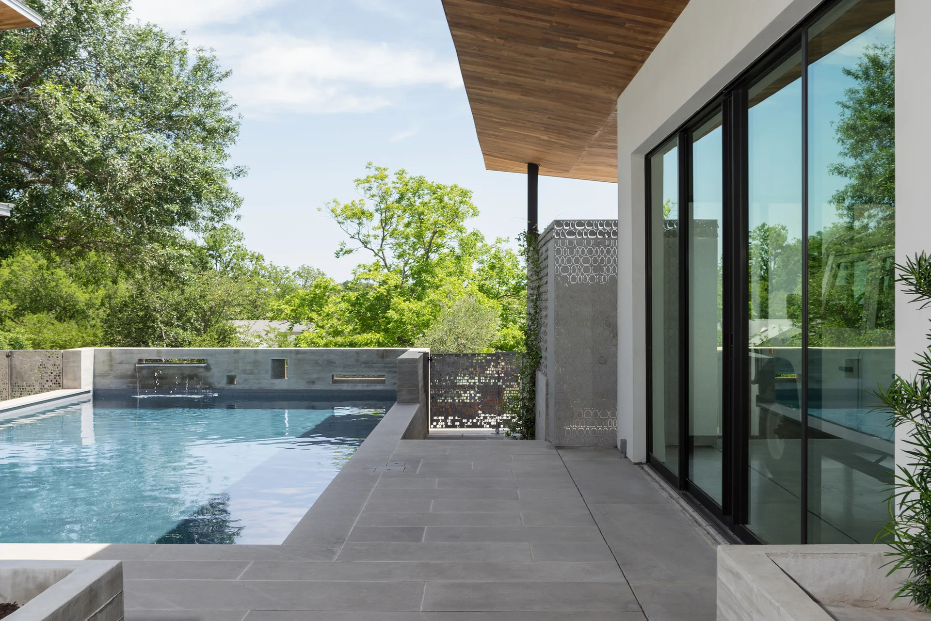 A modern outdoor swimming pool and concrete patio next to a house with large sliding glass doors, a wood plank overhang, and a geometric metal screen.