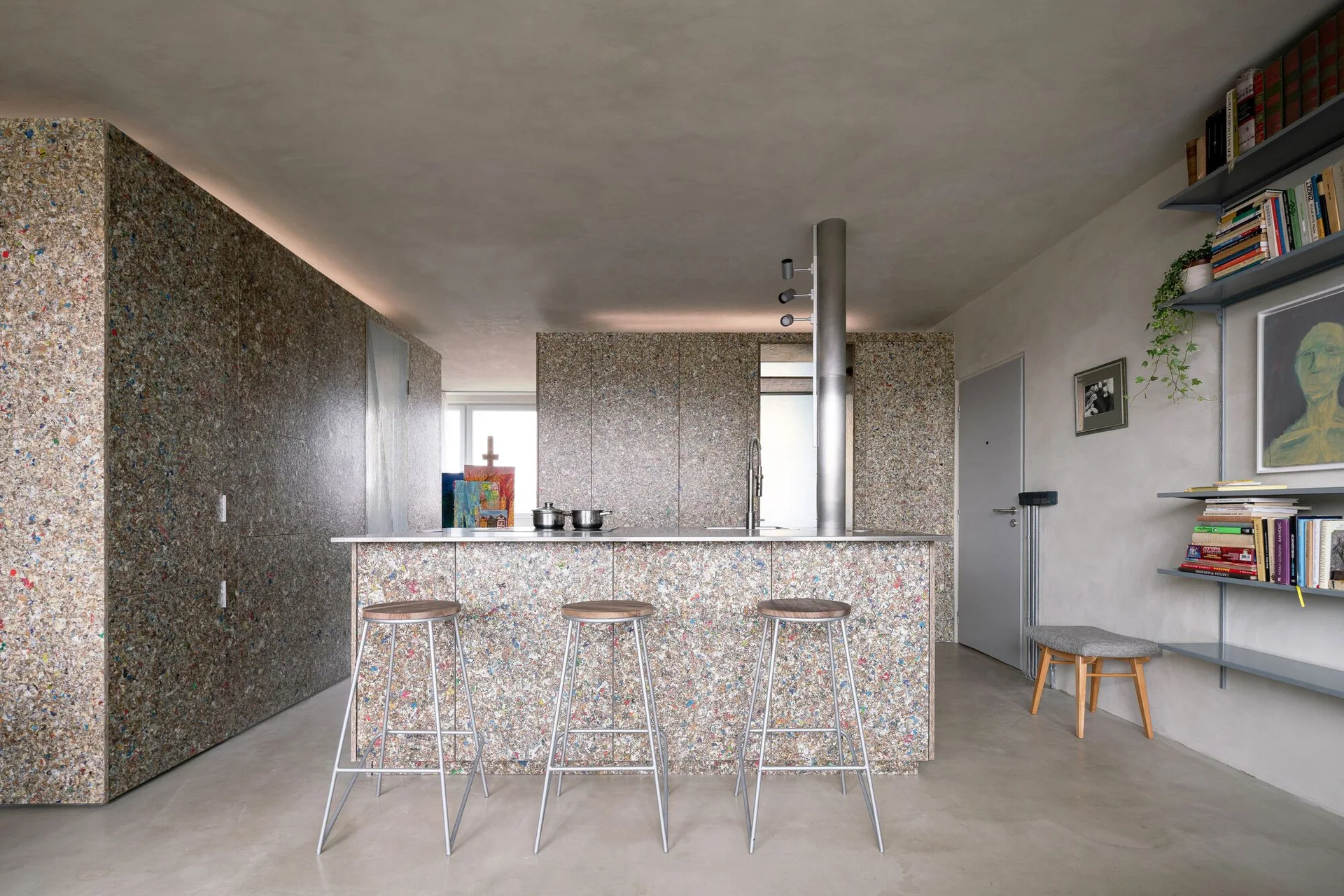 A modern kitchen featuring grey plaster walls, concrete floors, and cabinetry clad in a colorful speckled recycled composite material.
