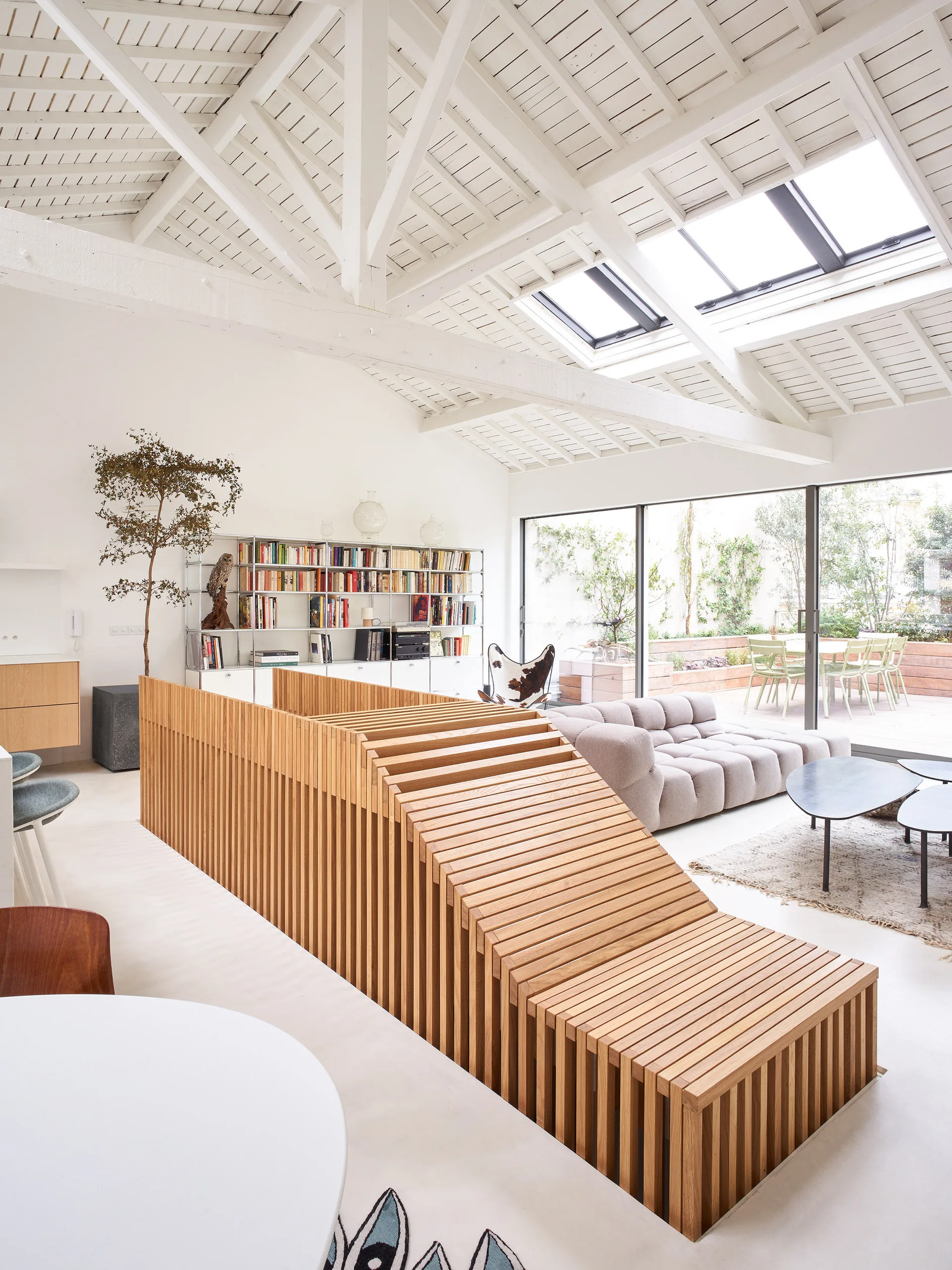 A bright loft living room features a unique slatted wood staircase structure, a tufted low sofa, and a high vaulted ceiling with skylights.