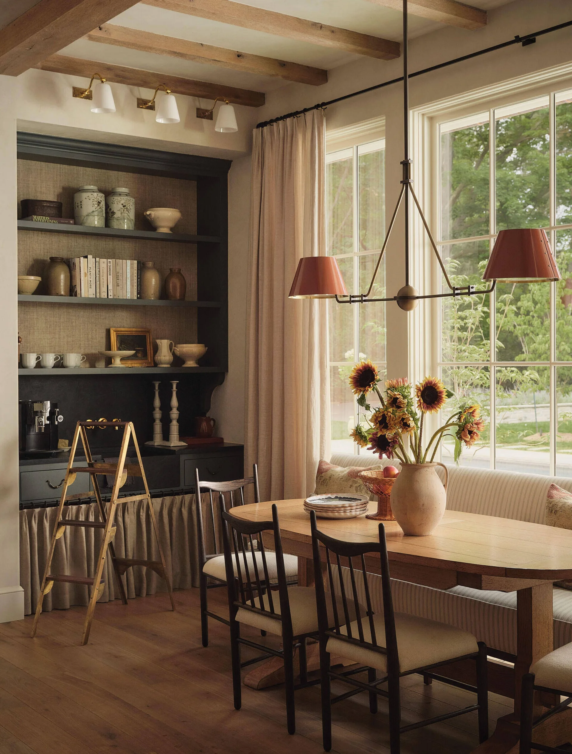 A rustic dining room features a wooden dining table, spindle-back chairs, a dark charcoal built-in cabinet, and a large window with banquette seating.