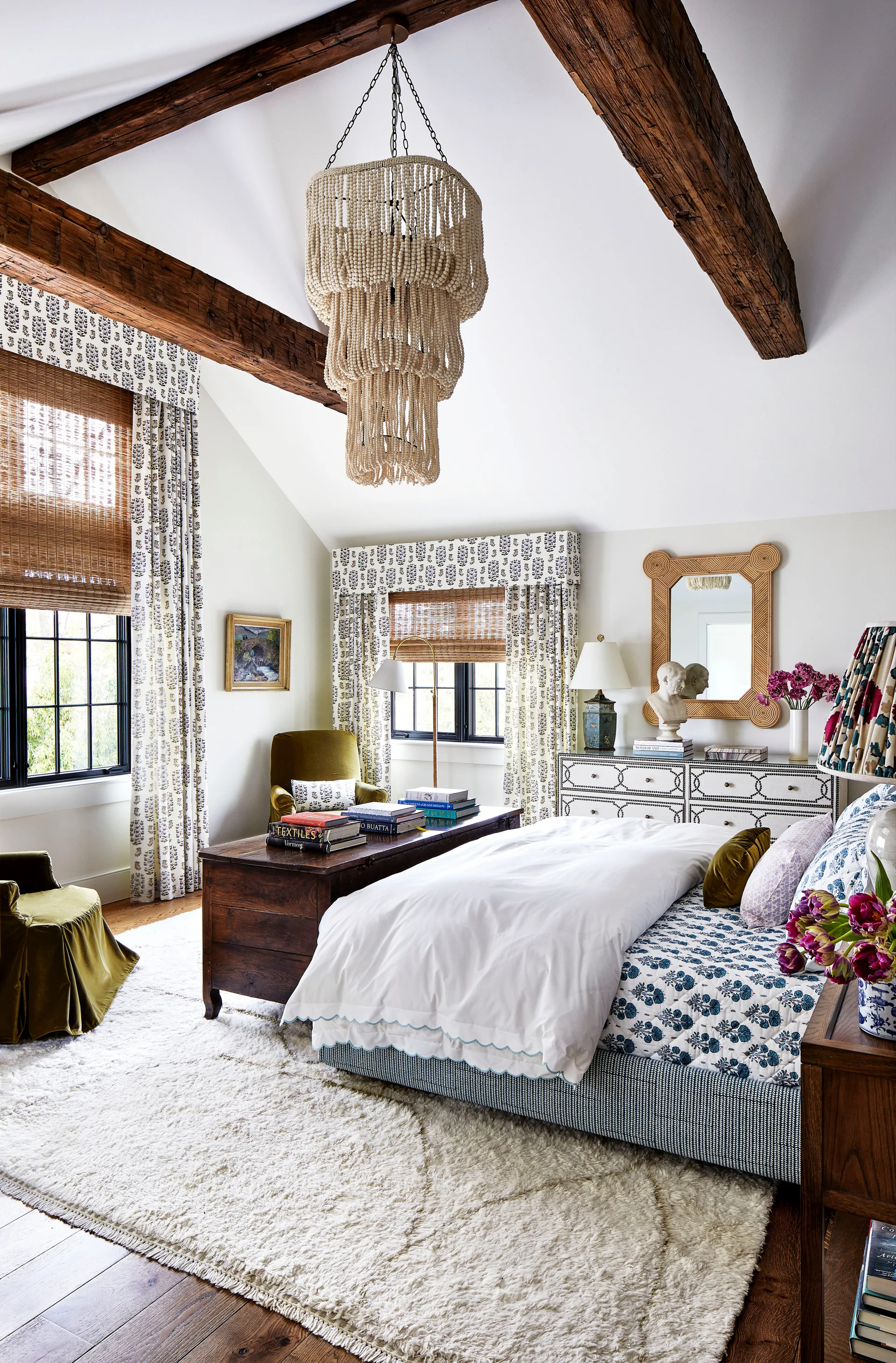 A well-lit bedroom features exposed dark wood ceiling beams, a large beaded chandelier, a bed with blue patterned linens, and a soft shag rug.