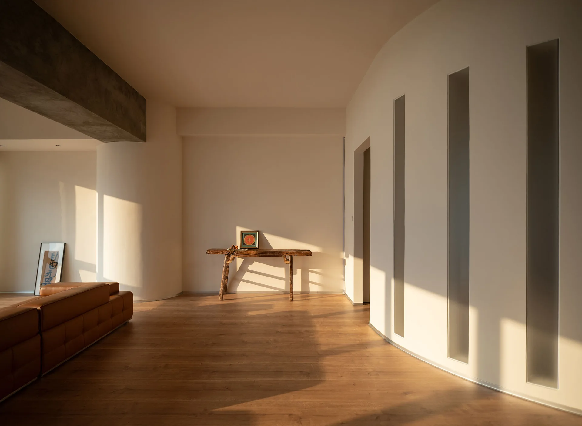 A minimalist living room features a curved warm beige wall with vertical glass slits, a rustic wooden console table, and wood floors in sunlight.
