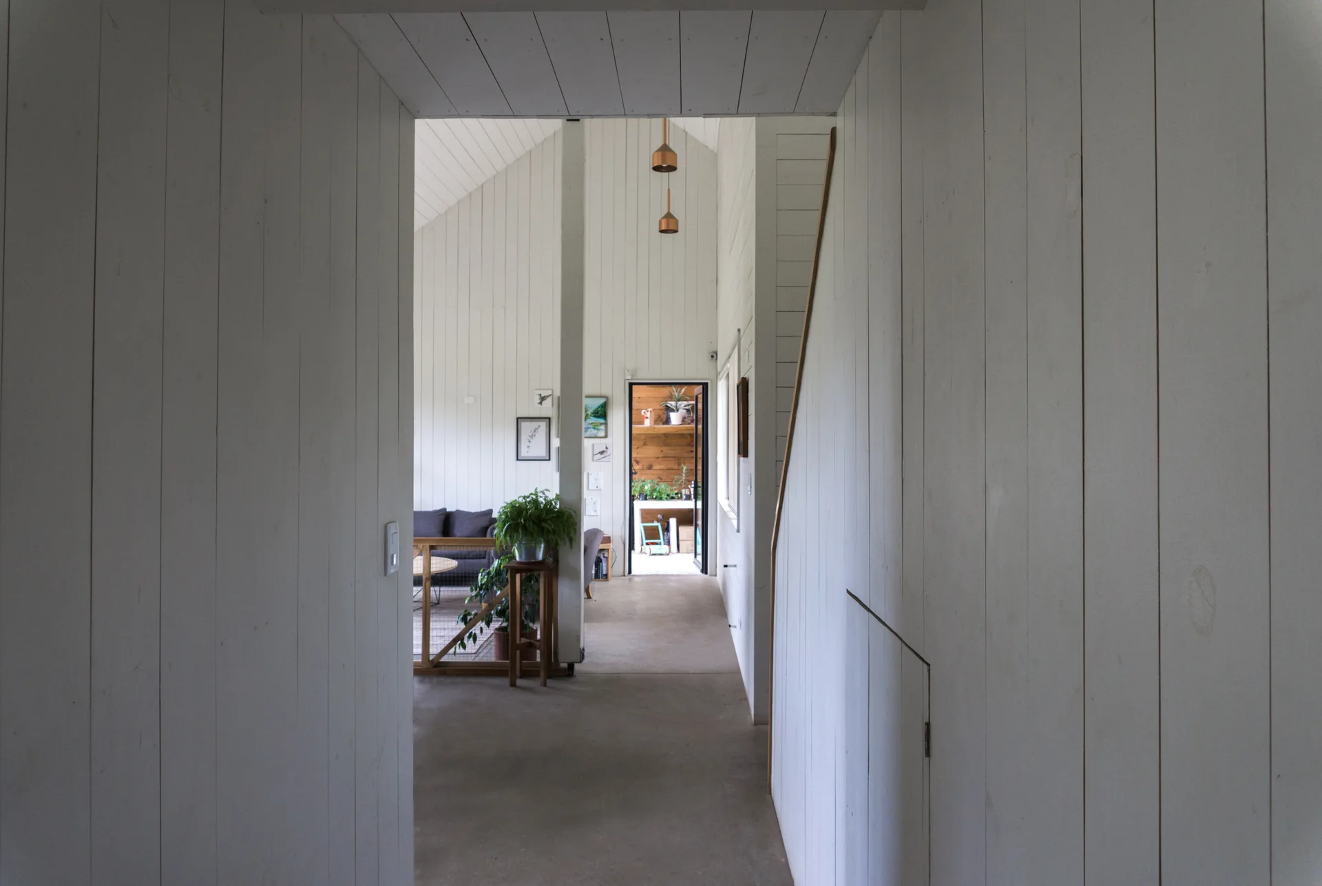 A view through a white vertical plank hallway leading into an open living room with a vaulted ceiling and copper pendant lights.