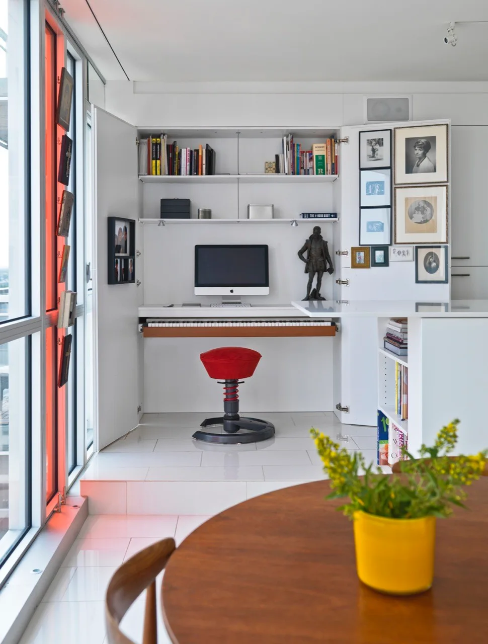 A white built-in home office features an integrated computer desk, a slide-out piano keyboard, open shelving, and a red ergonomic stool.