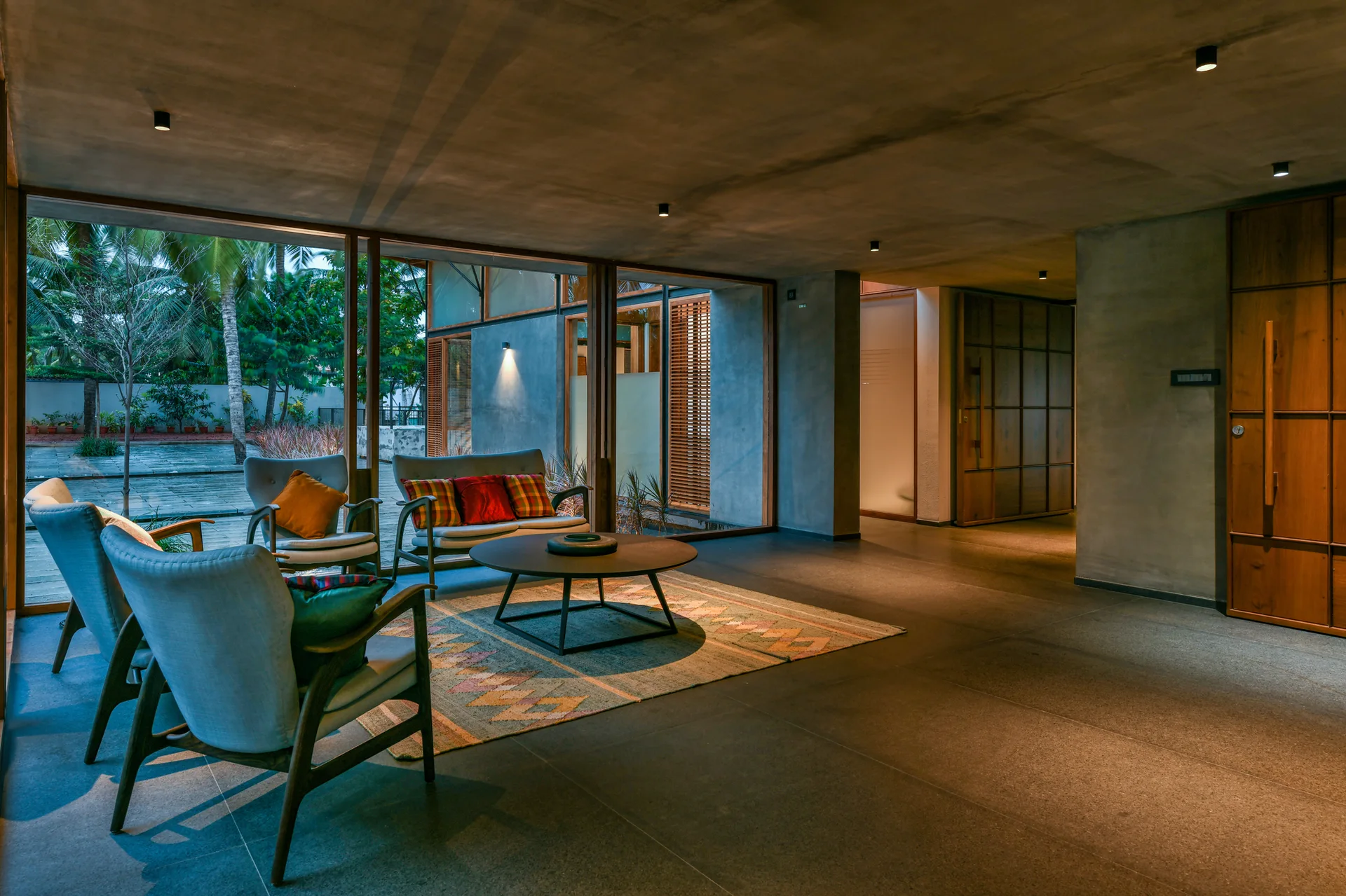 Mid-century modern armchairs and a round coffee table sit on a geometric rug in a living room with a concrete ceiling and large glass windows.