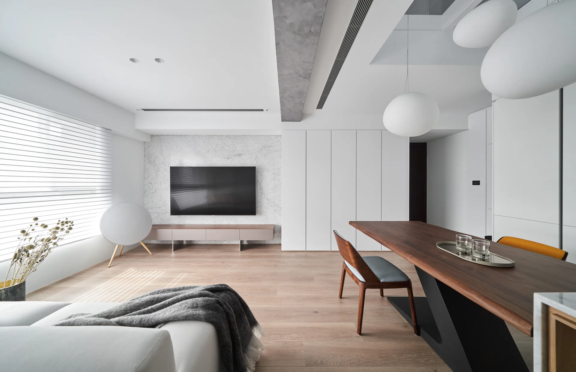 A minimalist living and dining space featuring a marble TV accent wall, built-in white cabinets, a dark wood table, and spherical pendant lights.