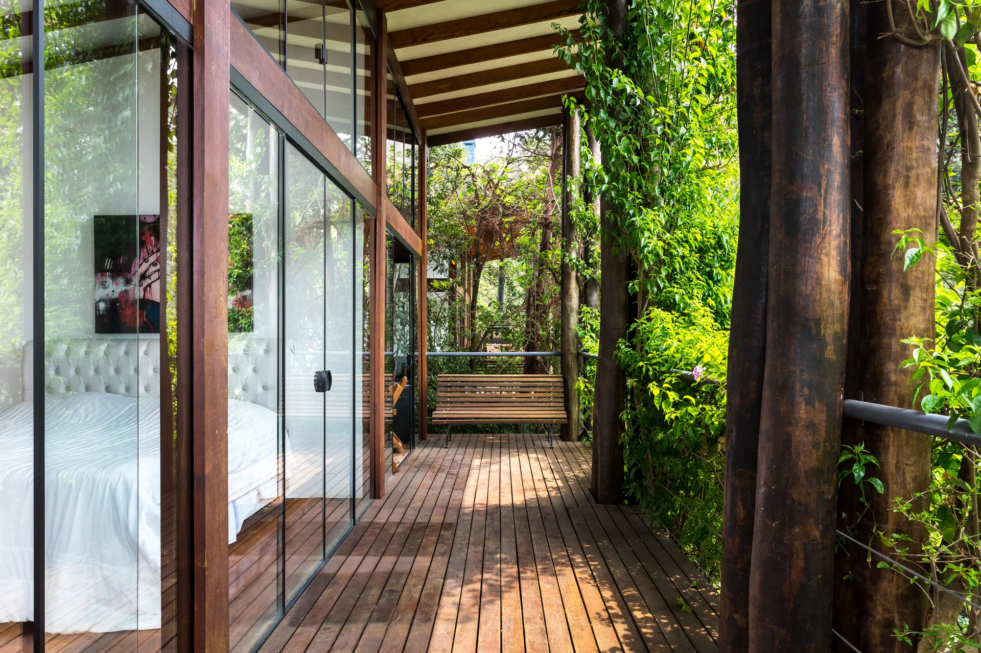 A wooden deck terrace with log pillars and a bench sits outside a bedroom with a white bed visible through floor-to-ceiling glass walls.