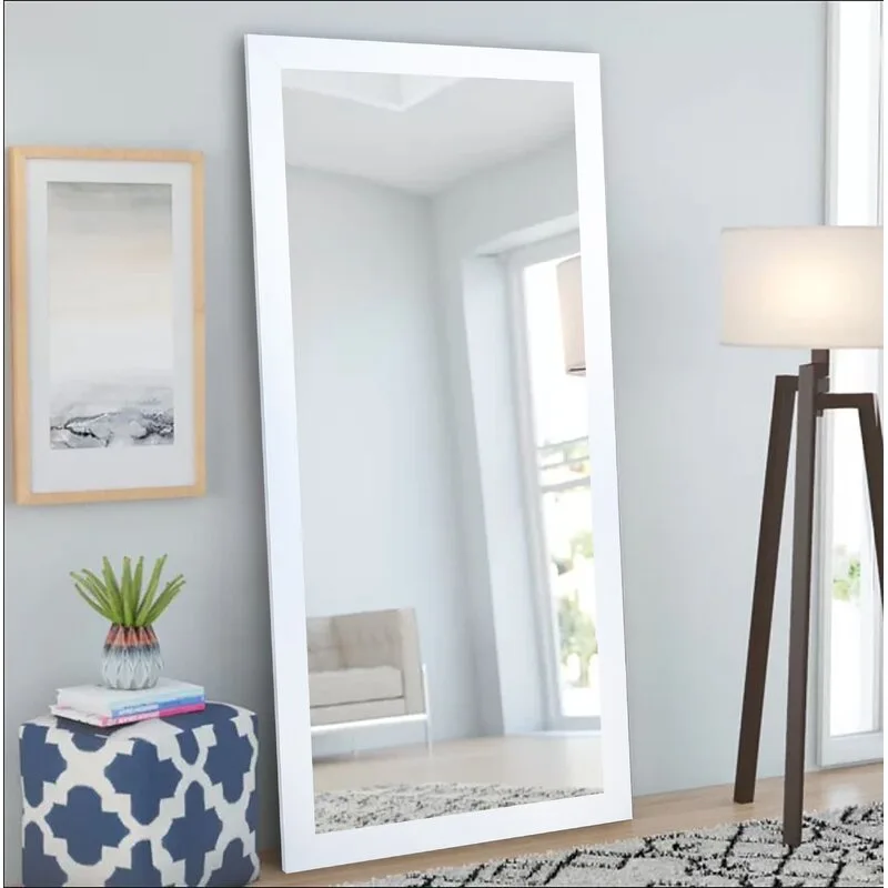 A large rectangular floor mirror with a thick white frame leans against a light grey wall next to a tripod lamp and a geometric blue ottoman.