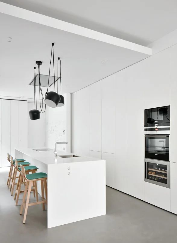 A minimalist white kitchen features a large island with teal cushioned bar stools under black pendant lights and a wall of handleless white cabinets.