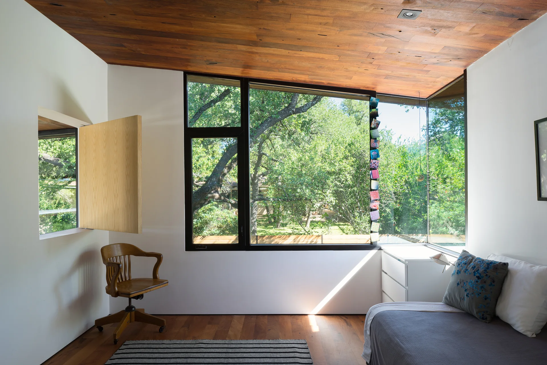 A sunlit bedroom features a warm wood ceiling, matching wood floors, white walls, and a corner window looking out at green trees.