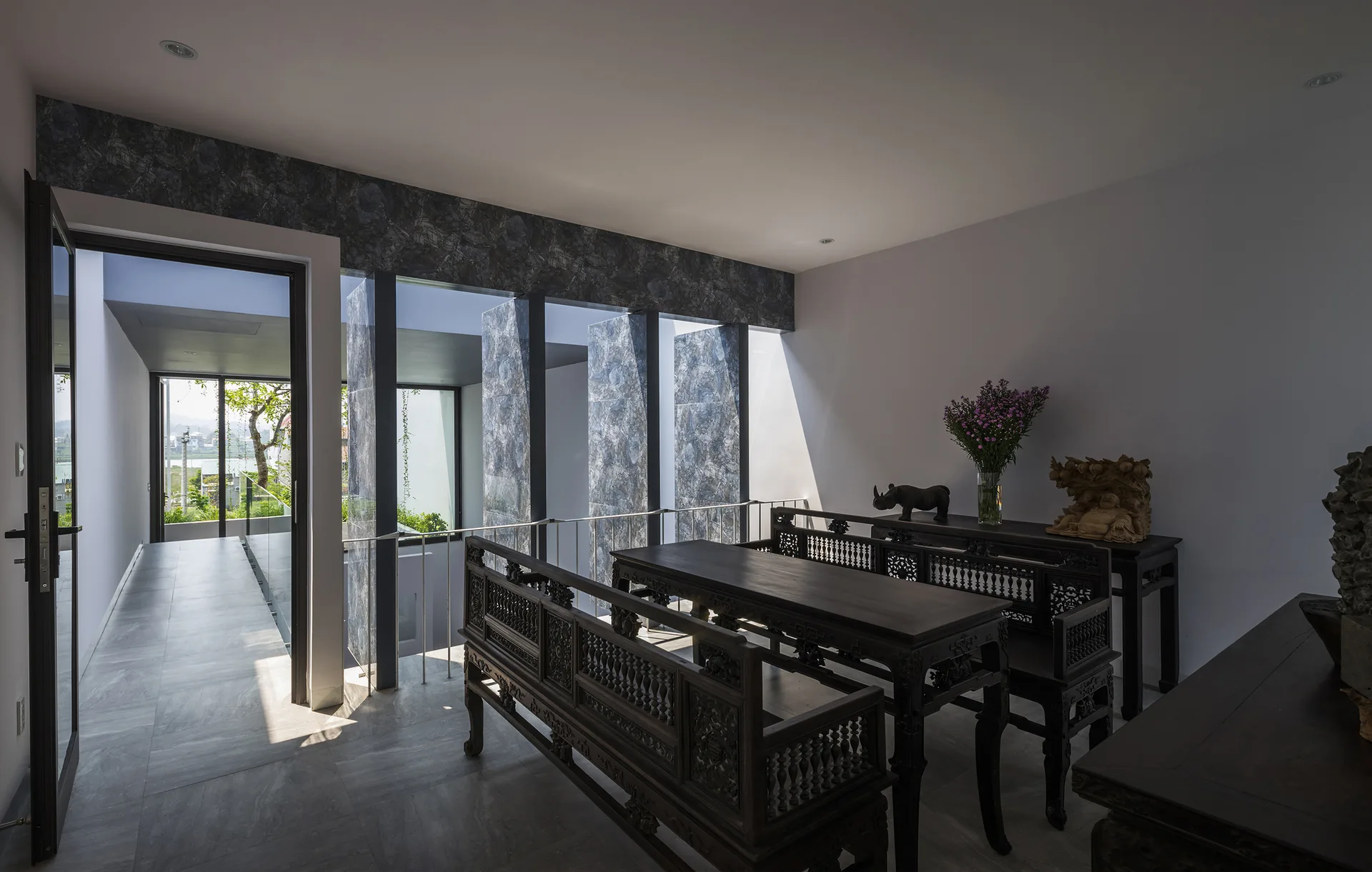 Ornate dark wood benches and tables stand in a modern room with gray stone tile flooring, white walls, and vertical windows with textured stone accents.