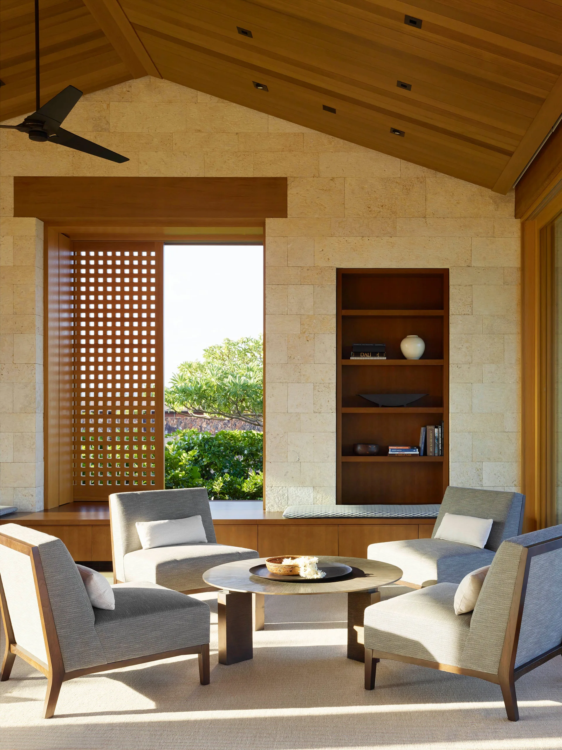 Four grey armchairs sit around a low round coffee table in a covered patio with block stone walls, a wood ceiling, and a sliding lattice window screen.