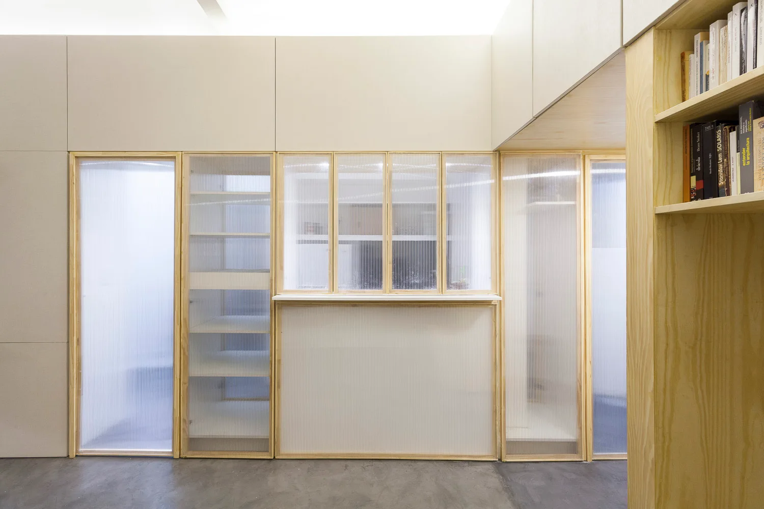 A minimalist interior partition wall features light wood framing, textured fluted glass panels, integrated shelving, and a grey concrete floor.